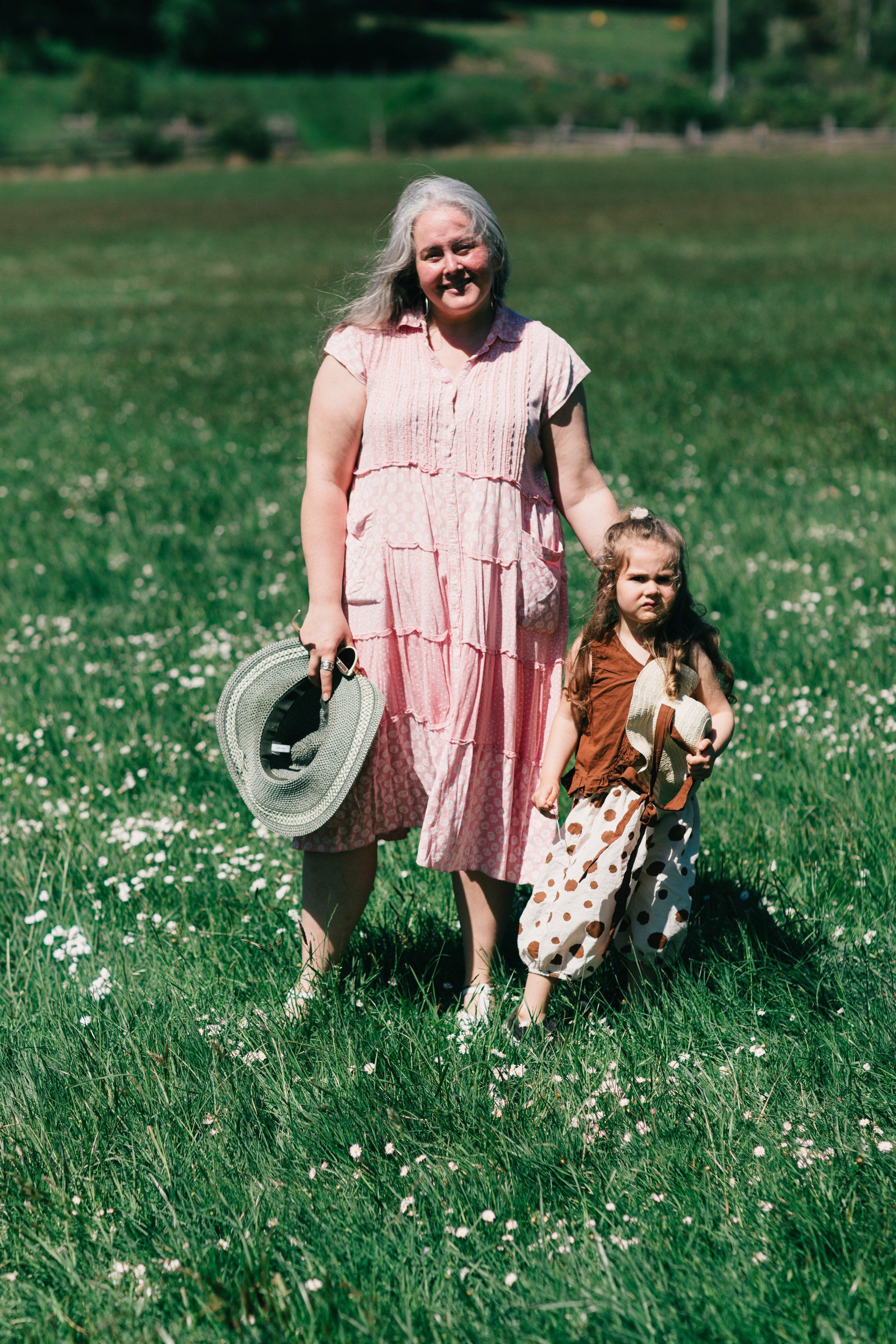 Woman in pink dress and child in brown jacket holding hands in blooming wildflower field, woman carrying wide-brimmed hat