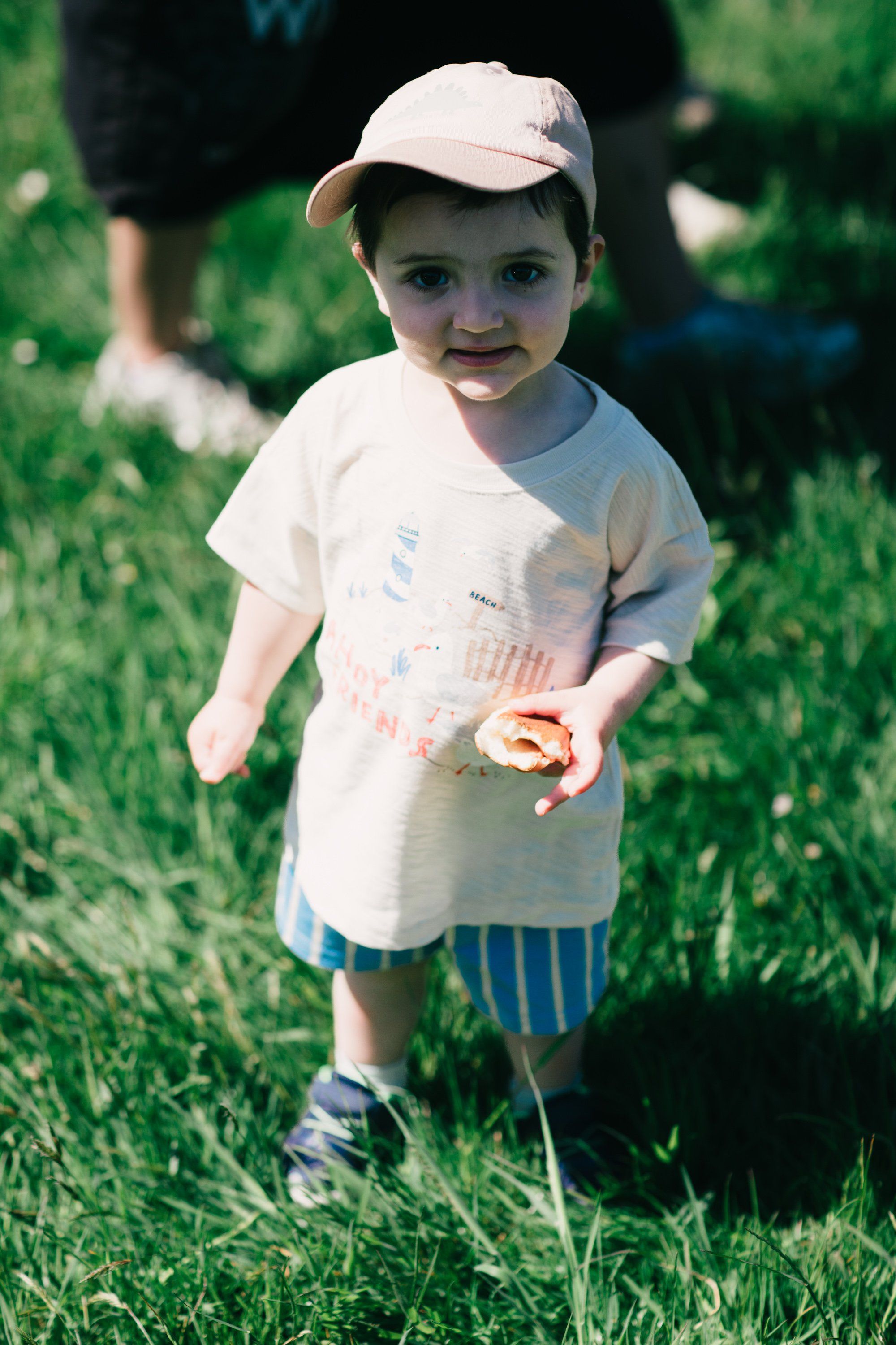 Toddler in white cap and shirt holding a donut while standing in sunlit grass at a farm