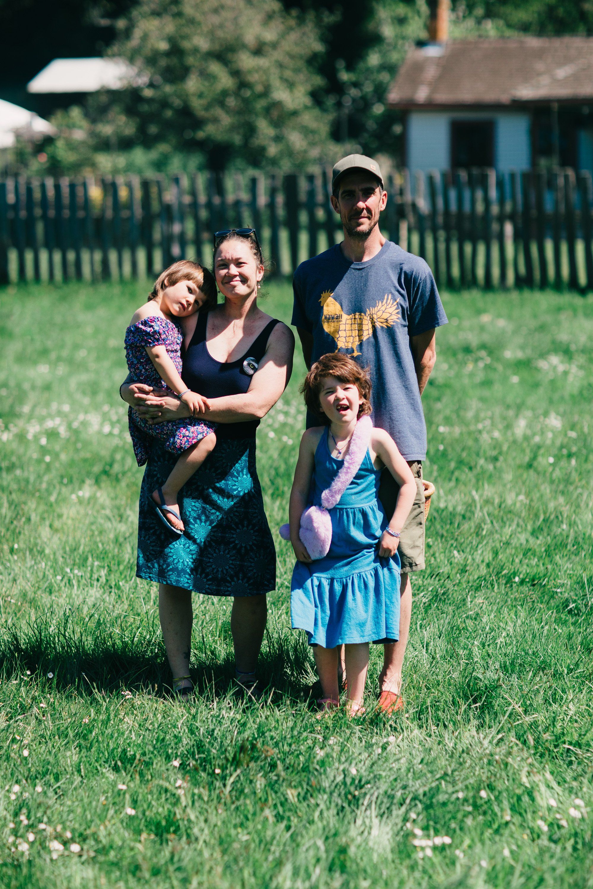 Family of four standing in a sunny grassy field with rural buildings and white fence behind them