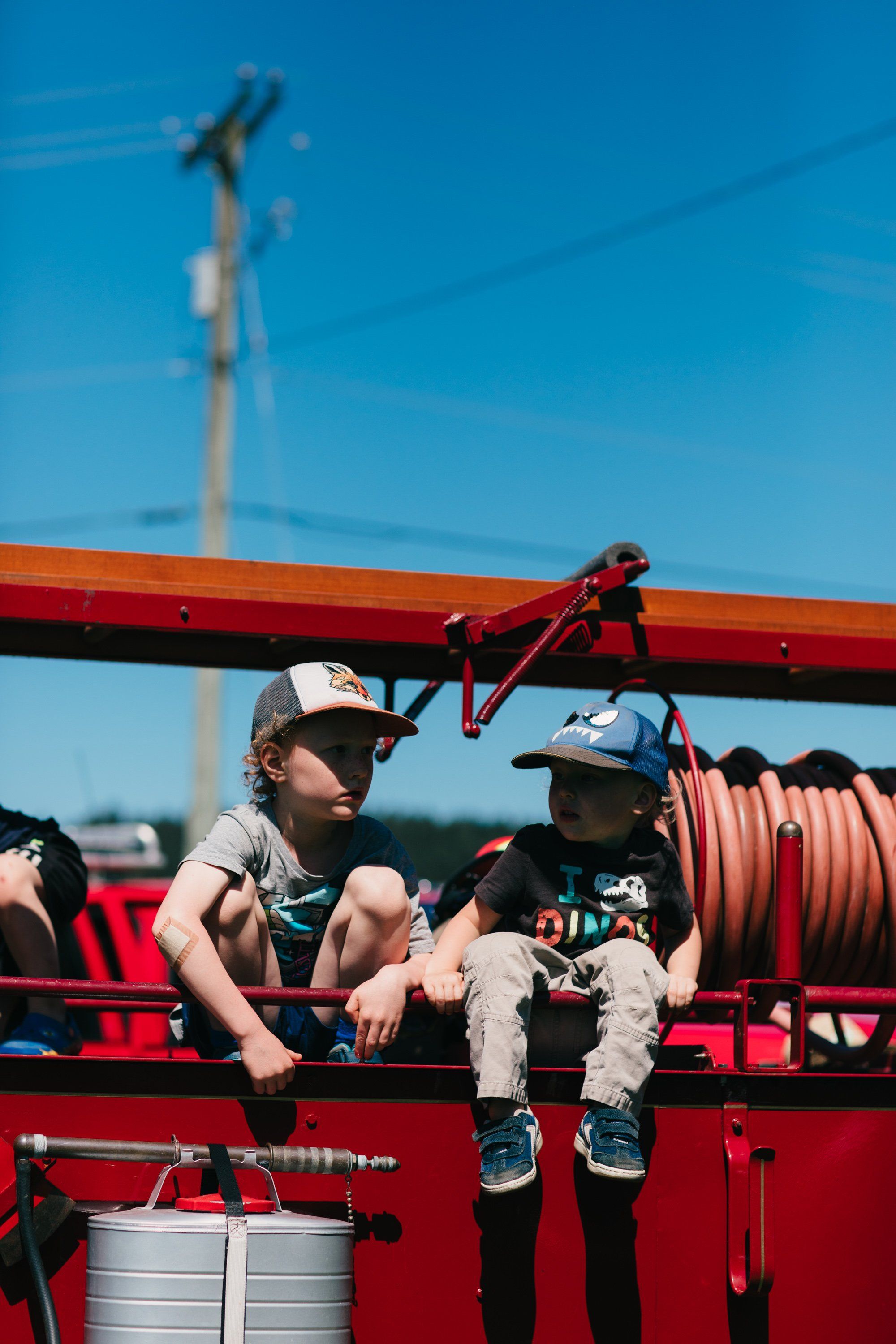 Two young children sit on a red farm implement against a bright blue sky, wearing caps and casual clothes