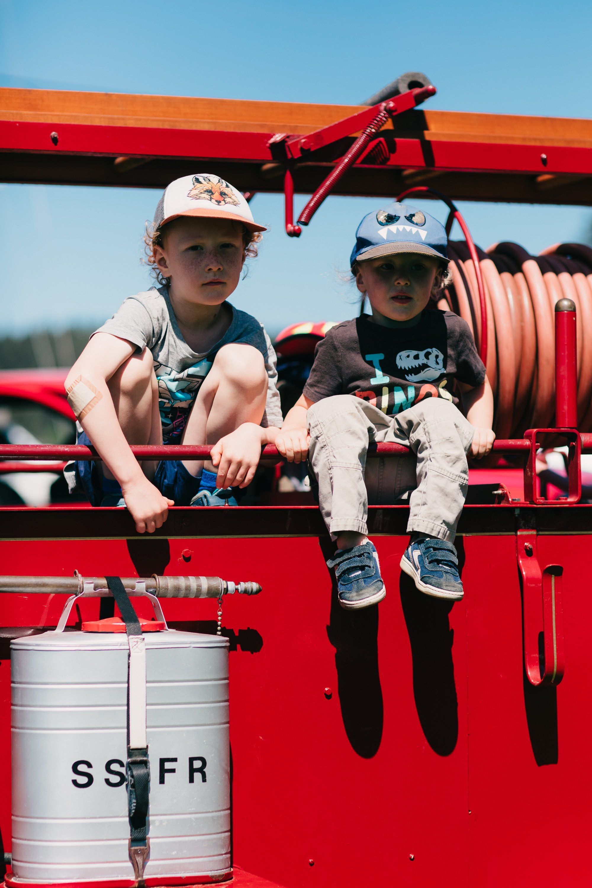 Two young children in white hard hats sit on a red farm vehicle under bright sunlight, legs dangling cheerfully over the side