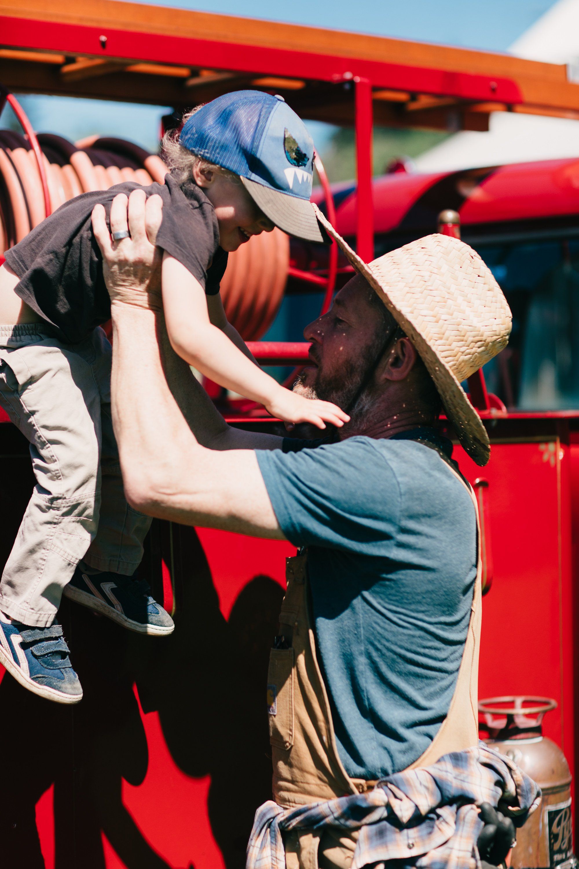 Adult lifting child toward red farm machinery under bright sunlight, wearing work clothes and hats