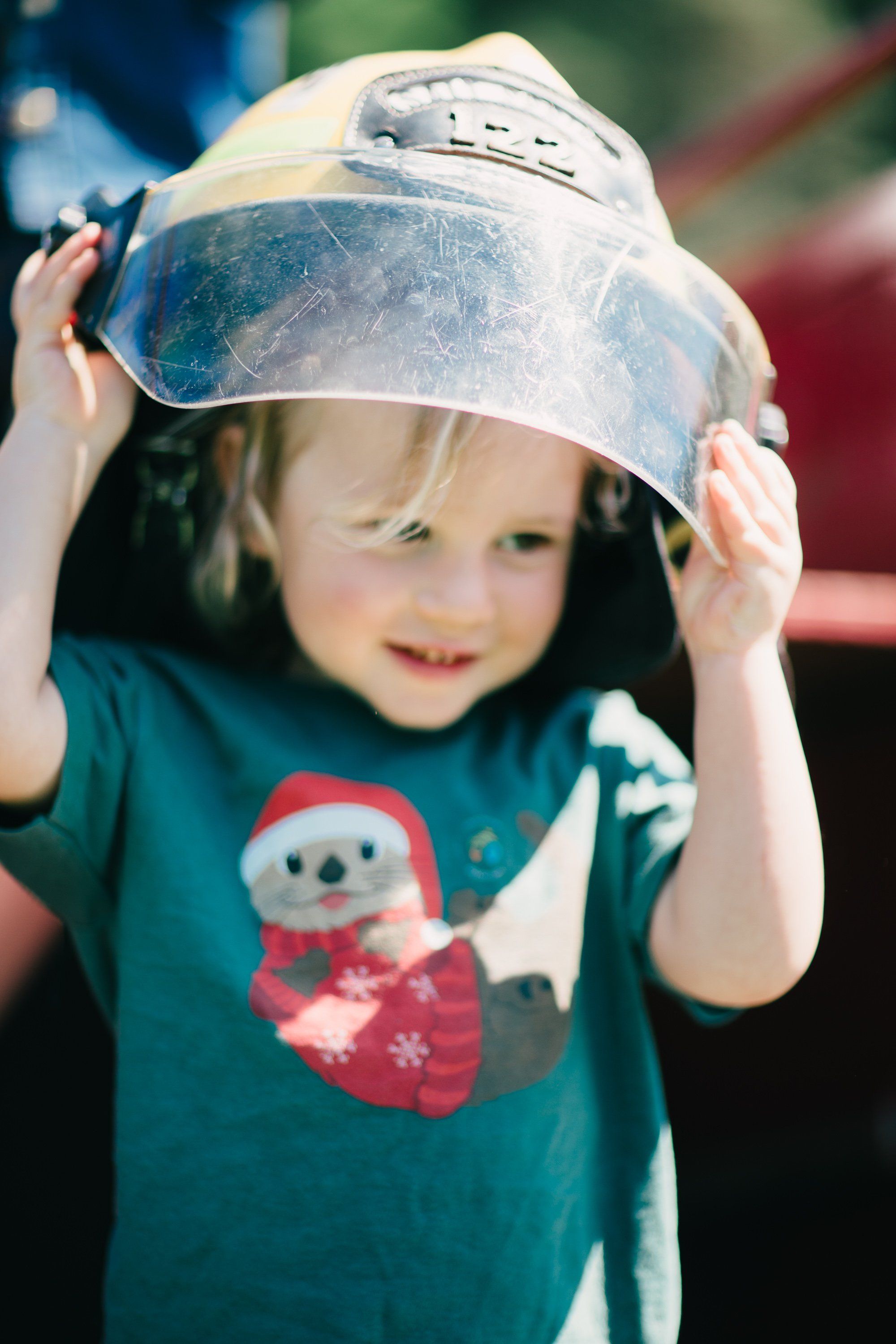 Young child in teal shirt with snowman design holds clear bowl over head, smiling at camera outdoors