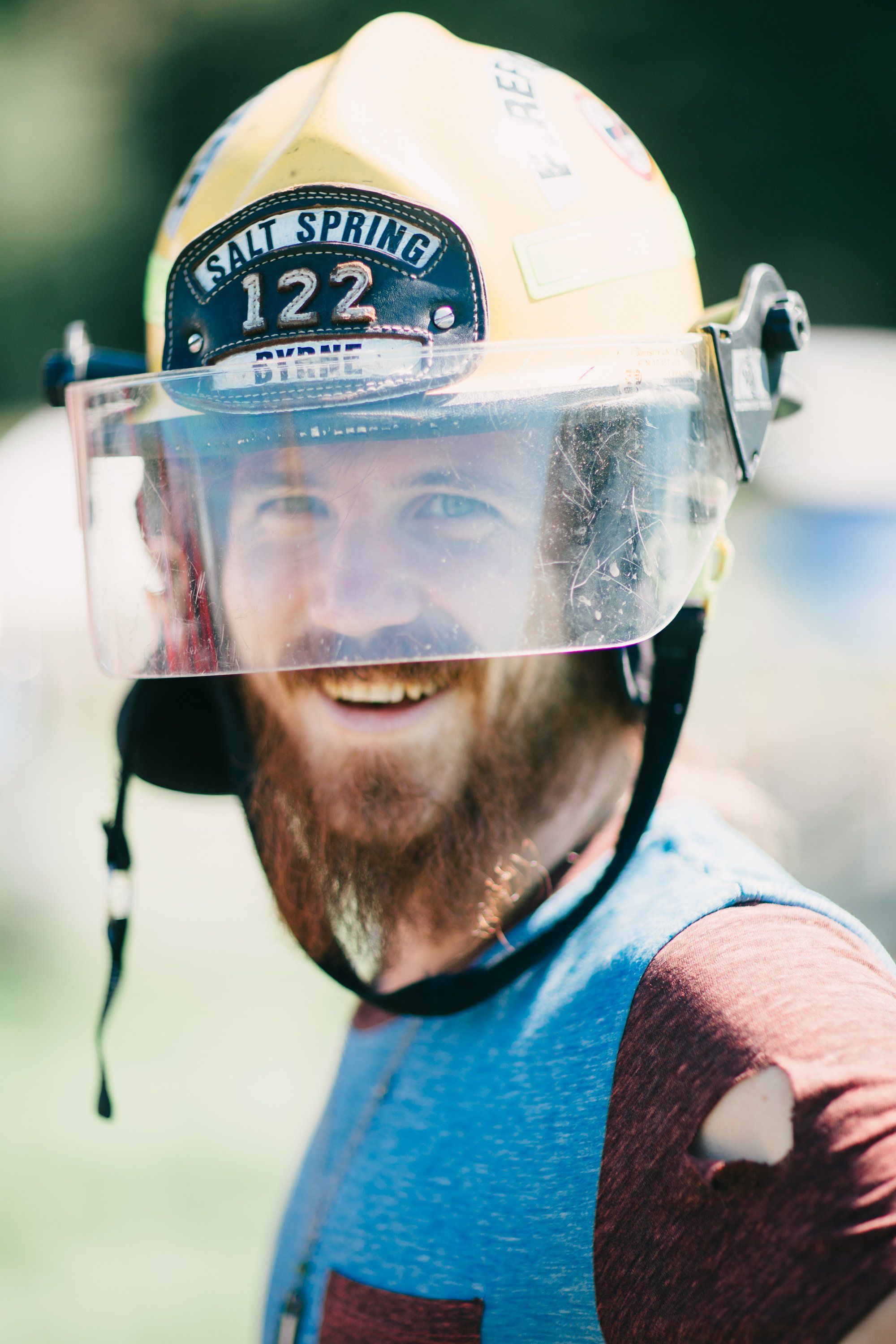 Bearded farmer in blue shirt wearing white firefighter helmet with clear face shield, smiling at camera during sunny day on farm fields