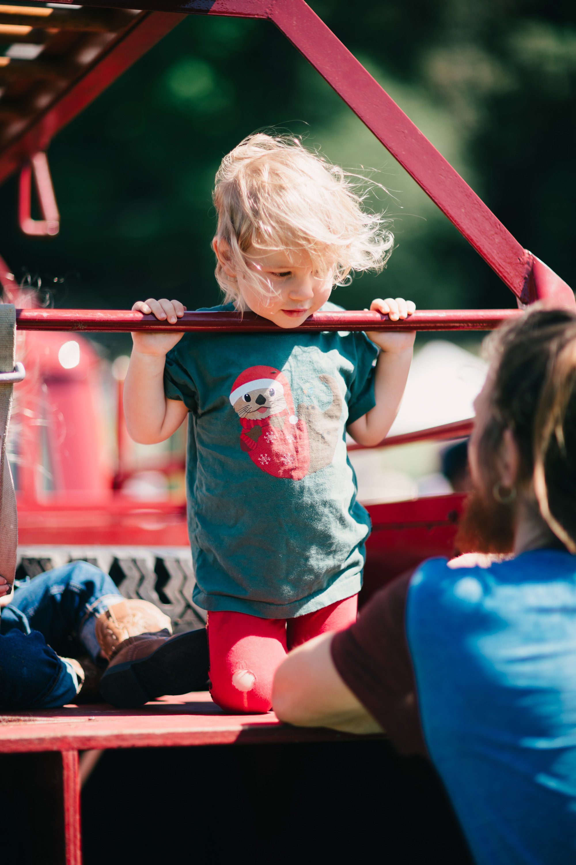 Blonde toddler in teal shirt stands on red farm equipment, bathed in golden sunlight, with blurred farmland background