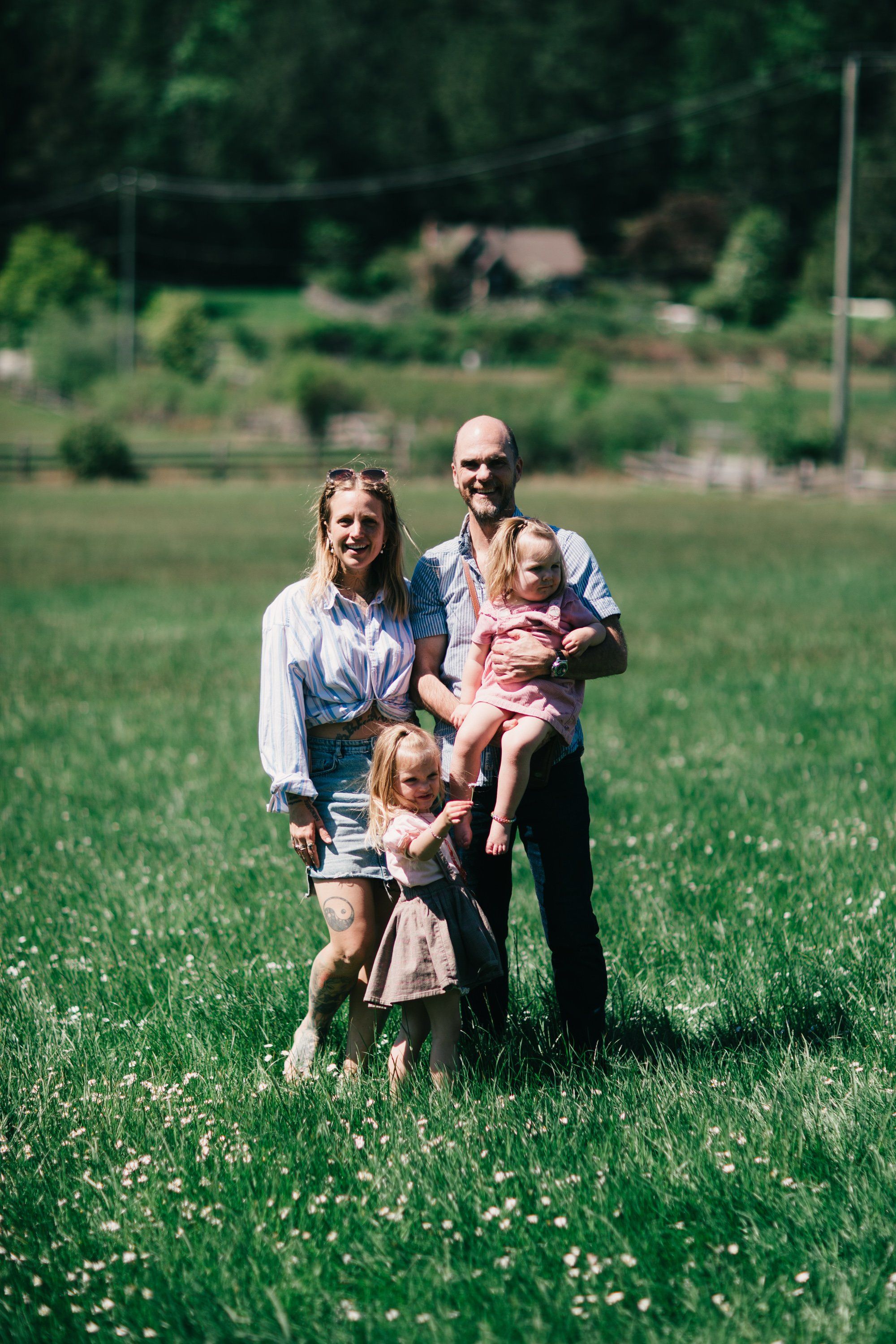 Family of four standing in a wildflower meadow with soft natural light and farm buildings visible in the blurred background