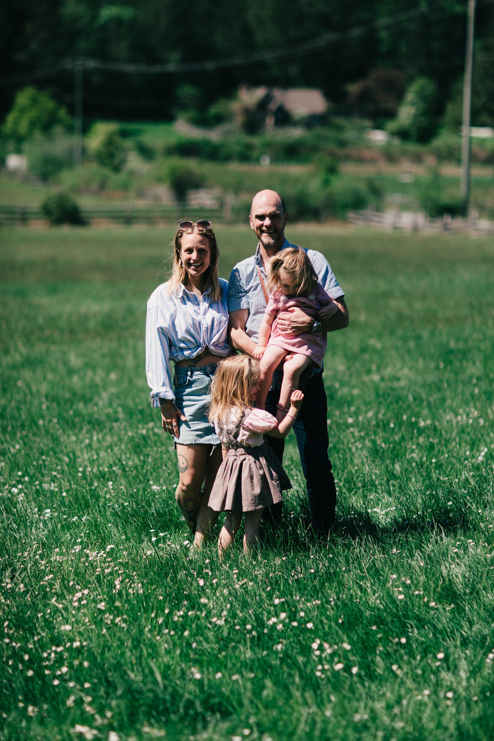 Family of three standing in a wildflower meadow with farm buildings visible in the soft-focused background