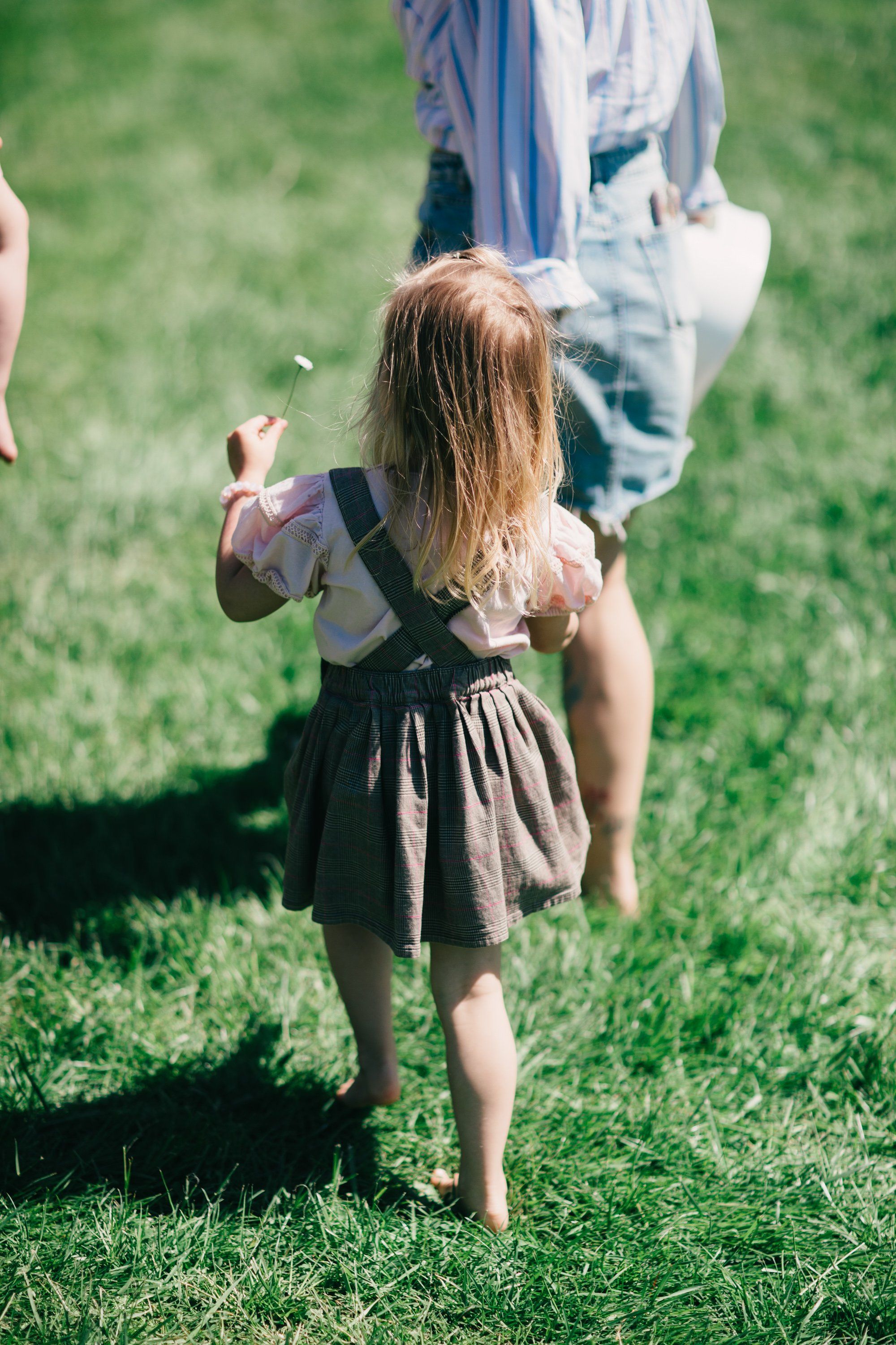 Toddler in a grey dress runs barefoot through green grass toward an adult in a blue shirt, dappled sunlight casting soft shadows