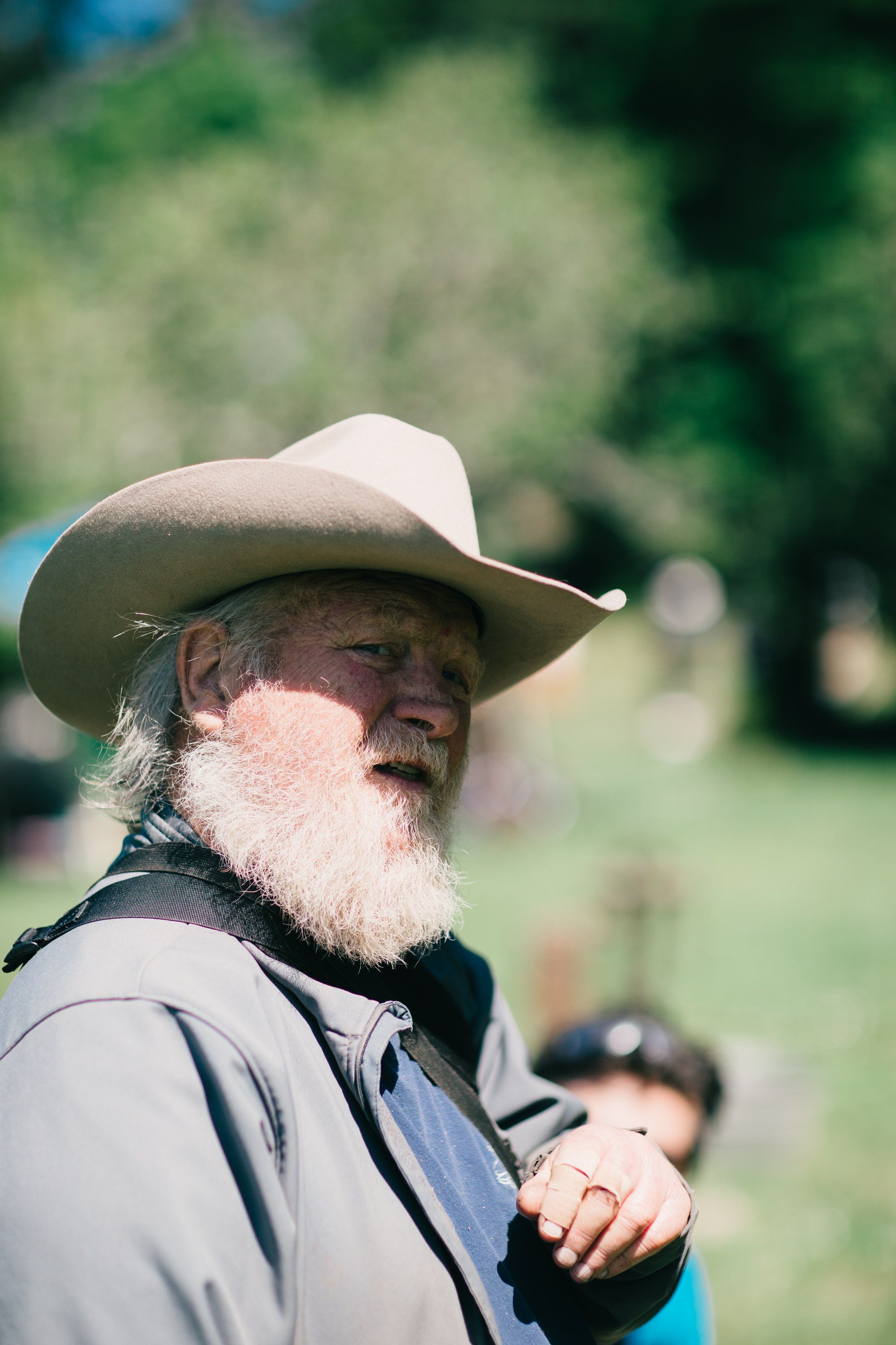 Bearded man in cream cowboy hat and gray jacket gazes across sunlit farm fields with soft green bokeh background