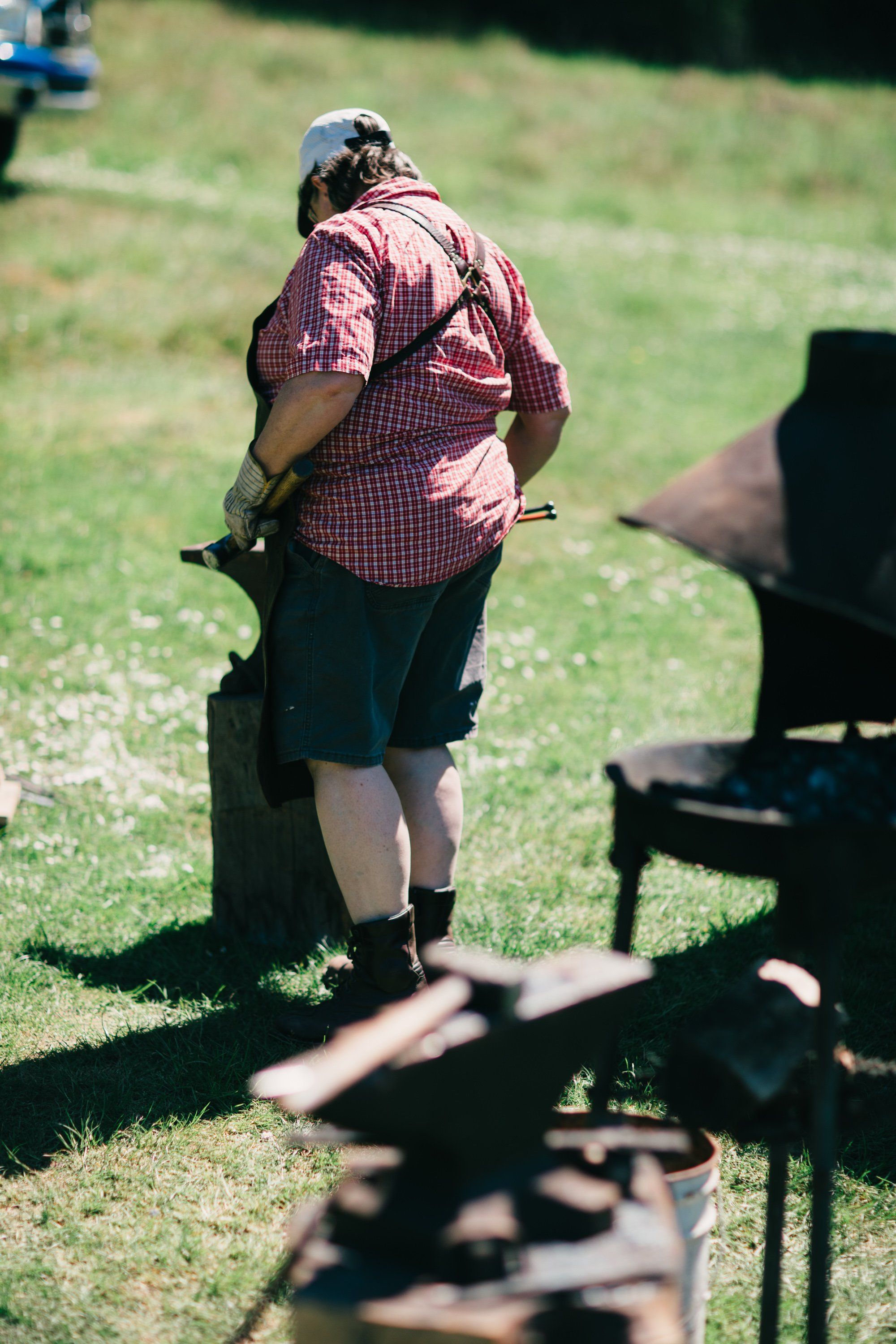 Man in red plaid shirt standing in bright sunlight on green grass at a farm, with farm equipment visible in foreground and background