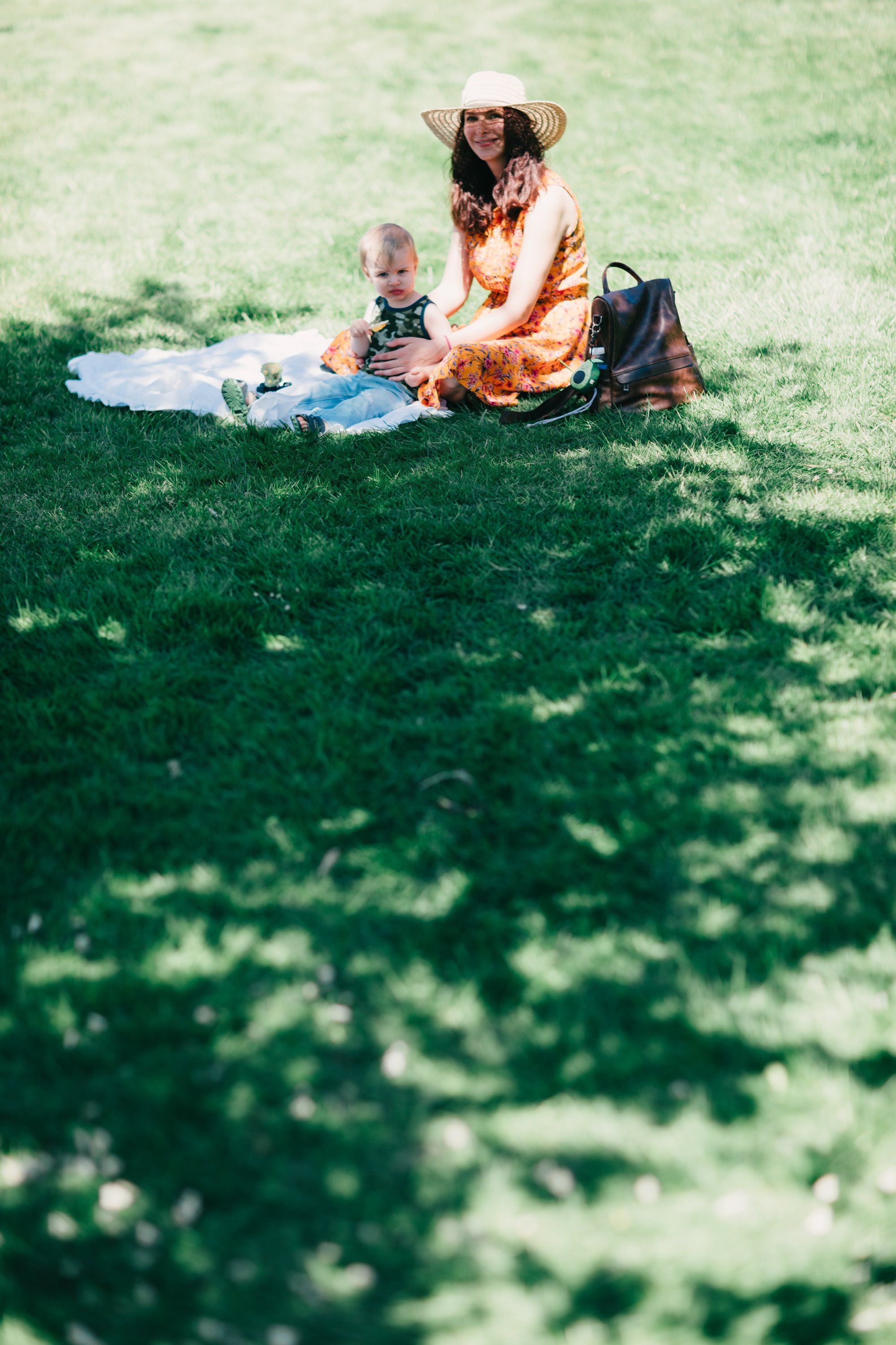 Woman in rust-colored shirt and wide-brimmed hat sits with toddler on sunlit grass, dappled shadows across the field