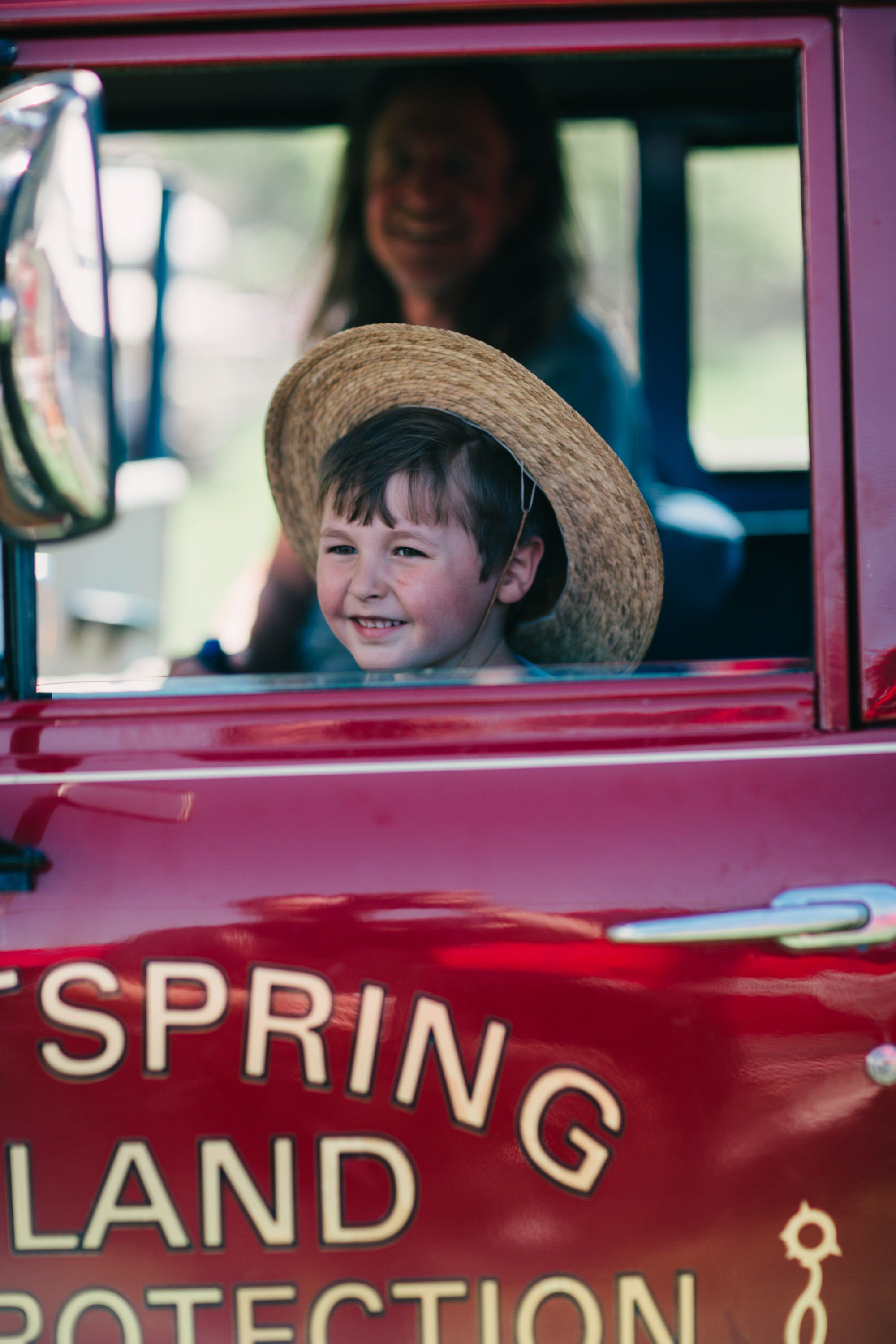 Young child in straw hat smiles from red farm vehicle window, adult visible behind in soft natural light