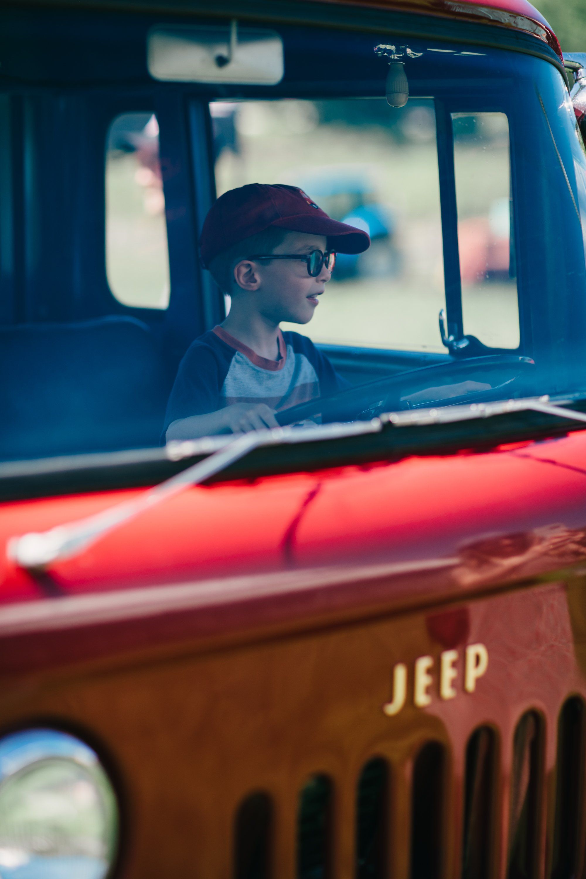 Child in baseball cap gazes out from rusty Jeep cabin, golden light catching the weathered farm vehicle's details