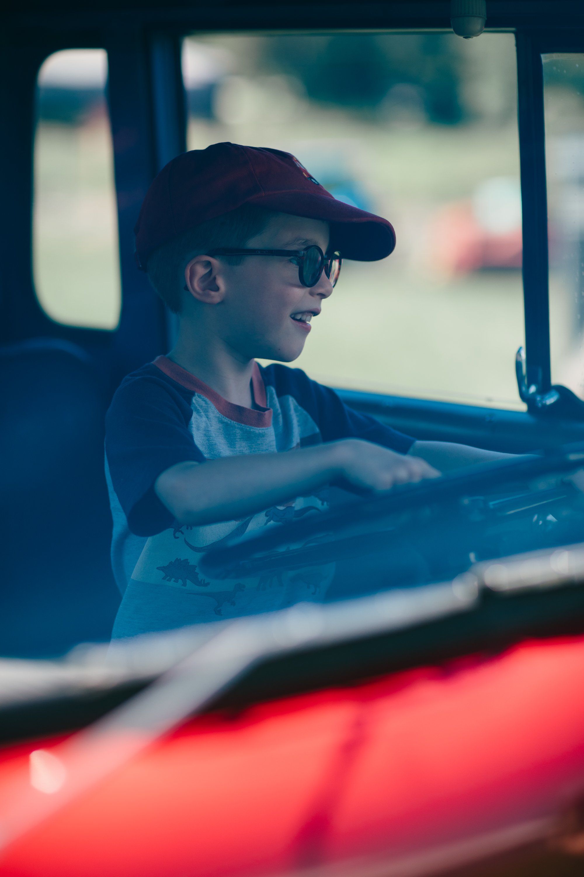 Young boy wearing glasses and a cap gazes out from inside a red farm vehicle on a sunny day