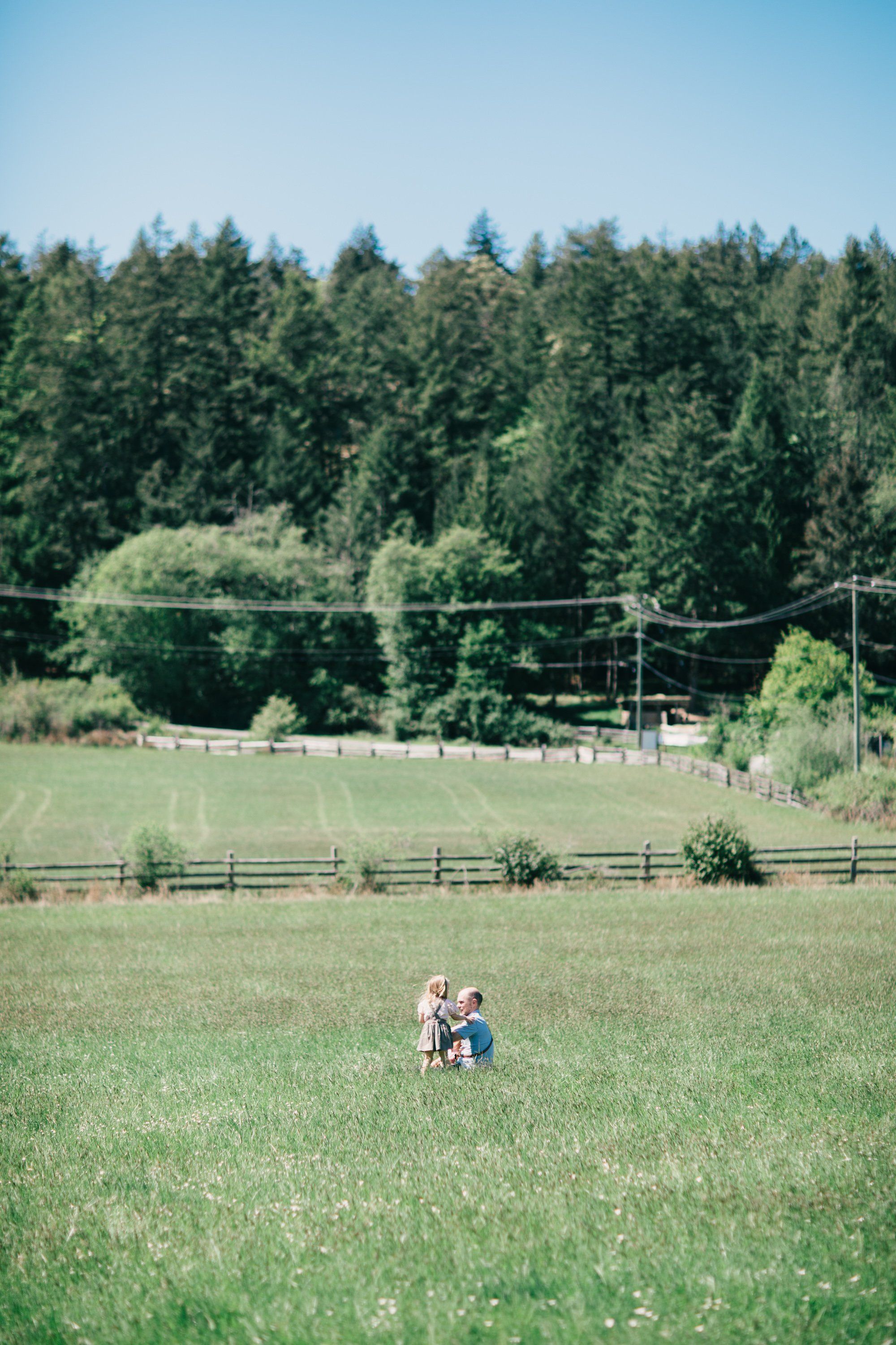Two children walk through a vast green pasture toward distant farm buildings and evergreen forest under clear blue sky