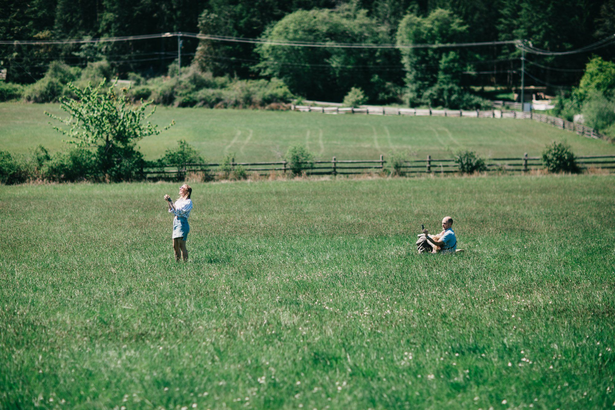 Two children run through a sunlit green field bordered by rustic wooden fences and dense forest