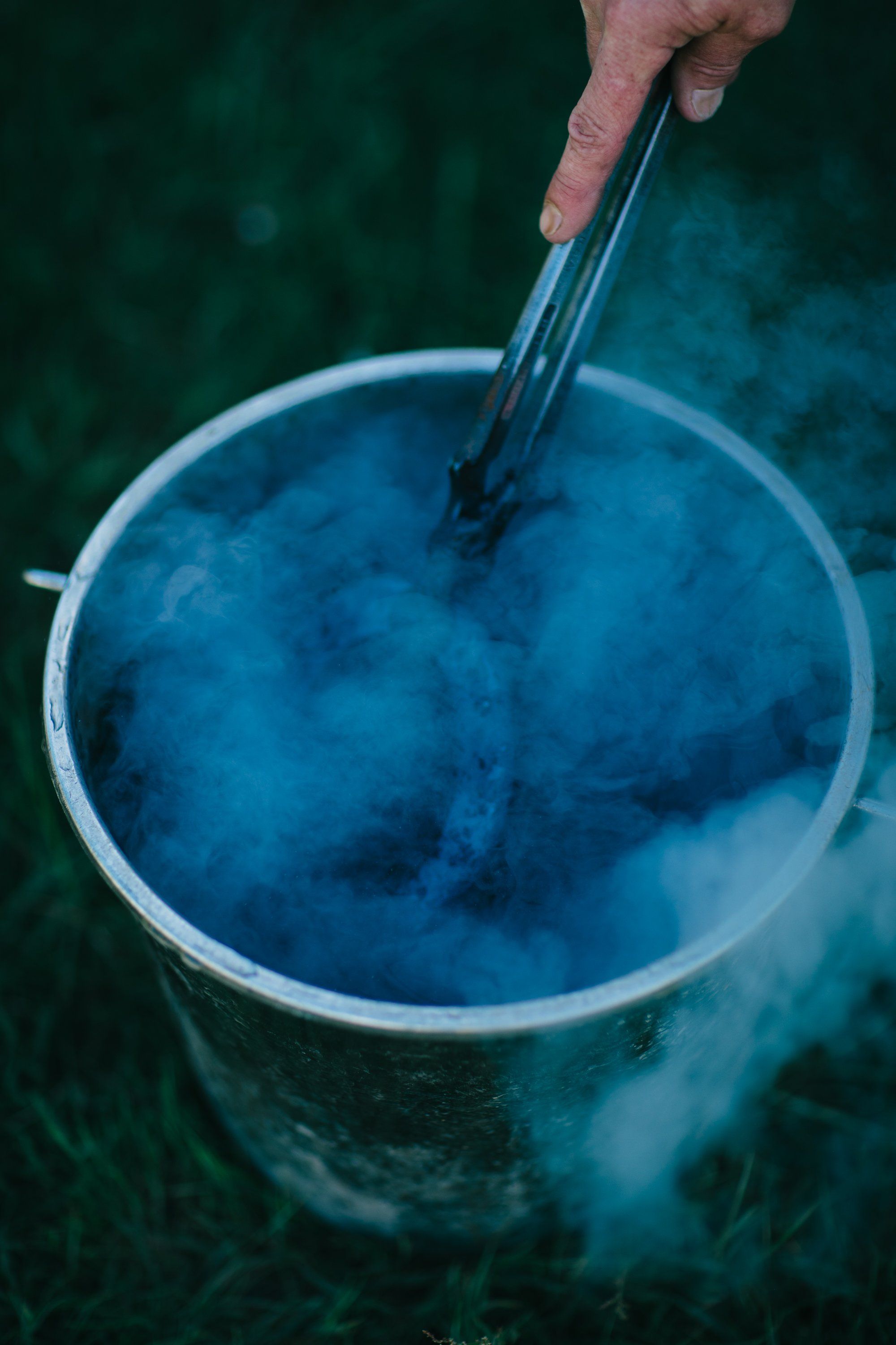 Hand stirring a bucket of deep indigo dye, steam rising against a dark forest backdrop