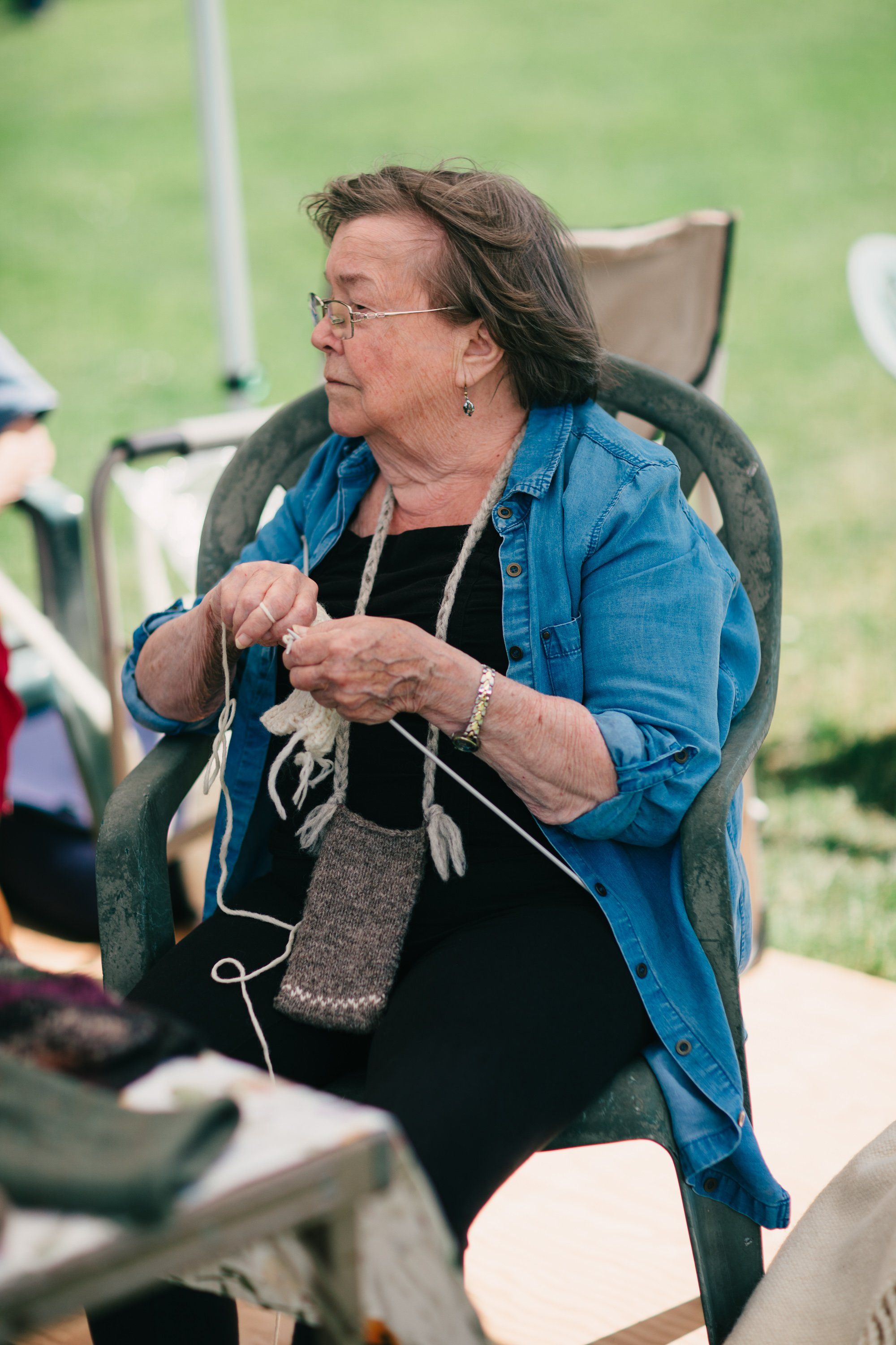 Woman knitting outdoors at Ruckle Farm, wearing glasses and denim shirt, seated in camping chair with green field behind her