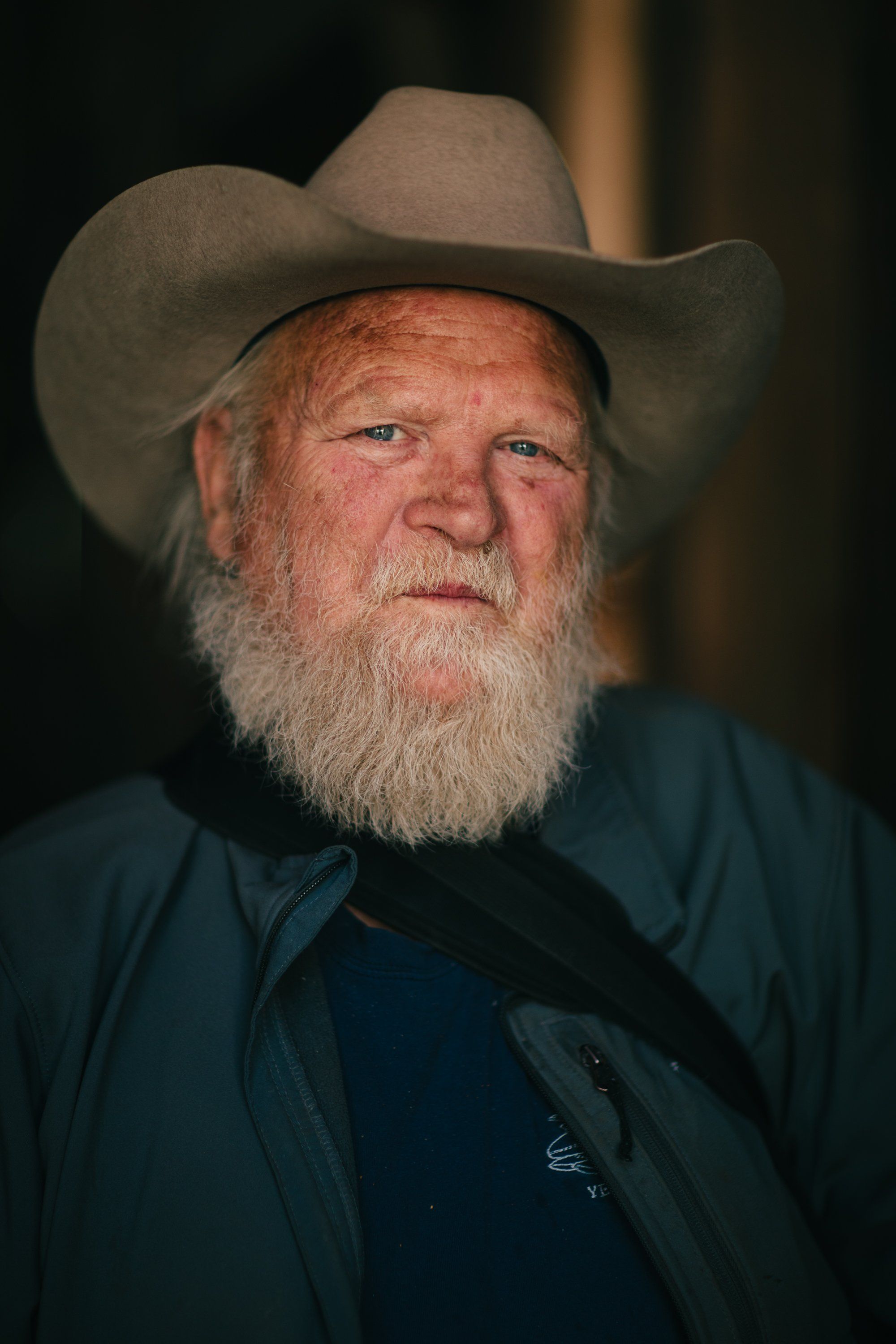 Weathered man with full white beard wearing a tan cowboy hat and dark jacket, looking upward with contemplative expression