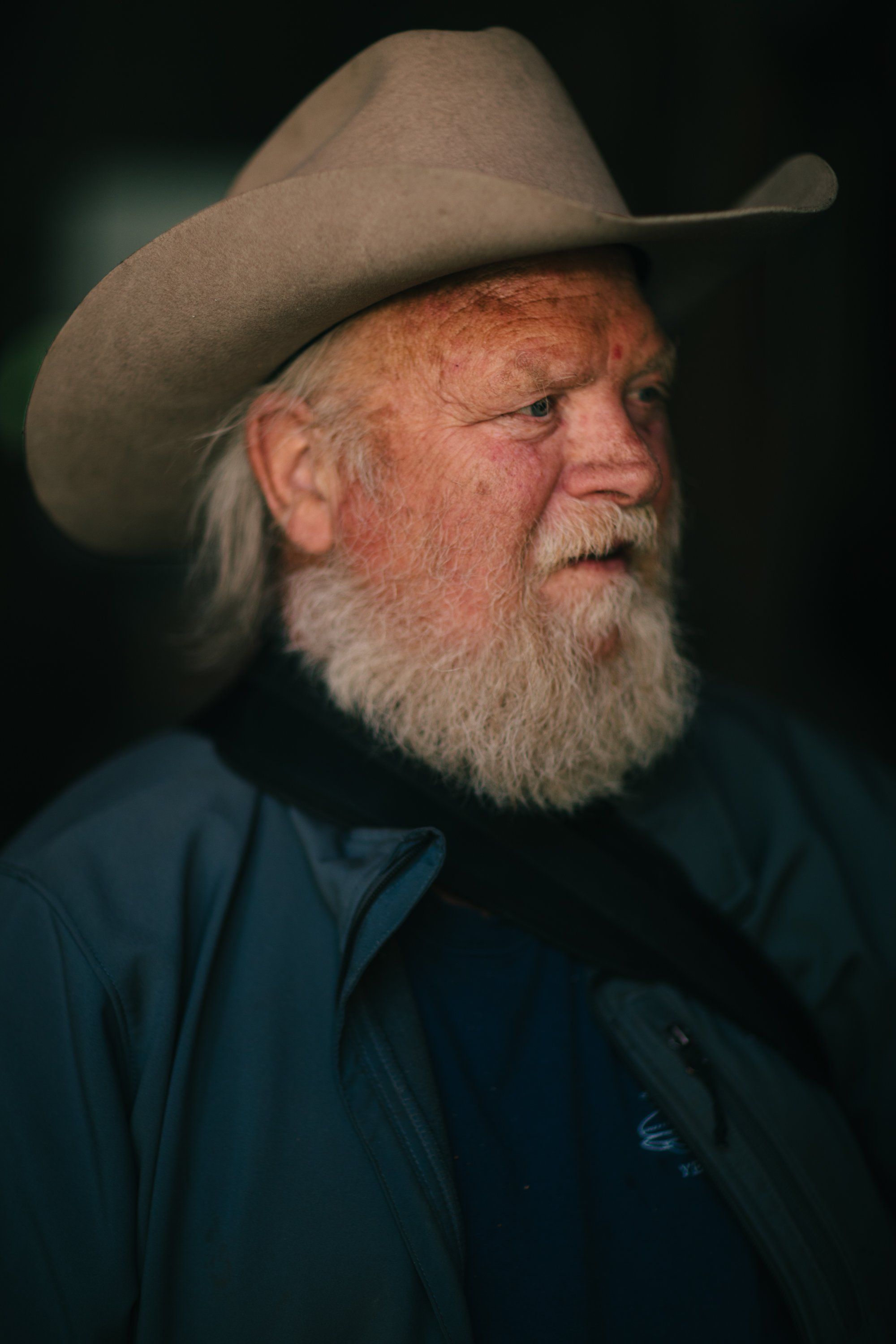 Weathered older man with long white beard wearing a brown cowboy hat and dark jacket, photographed in profile against a dark background