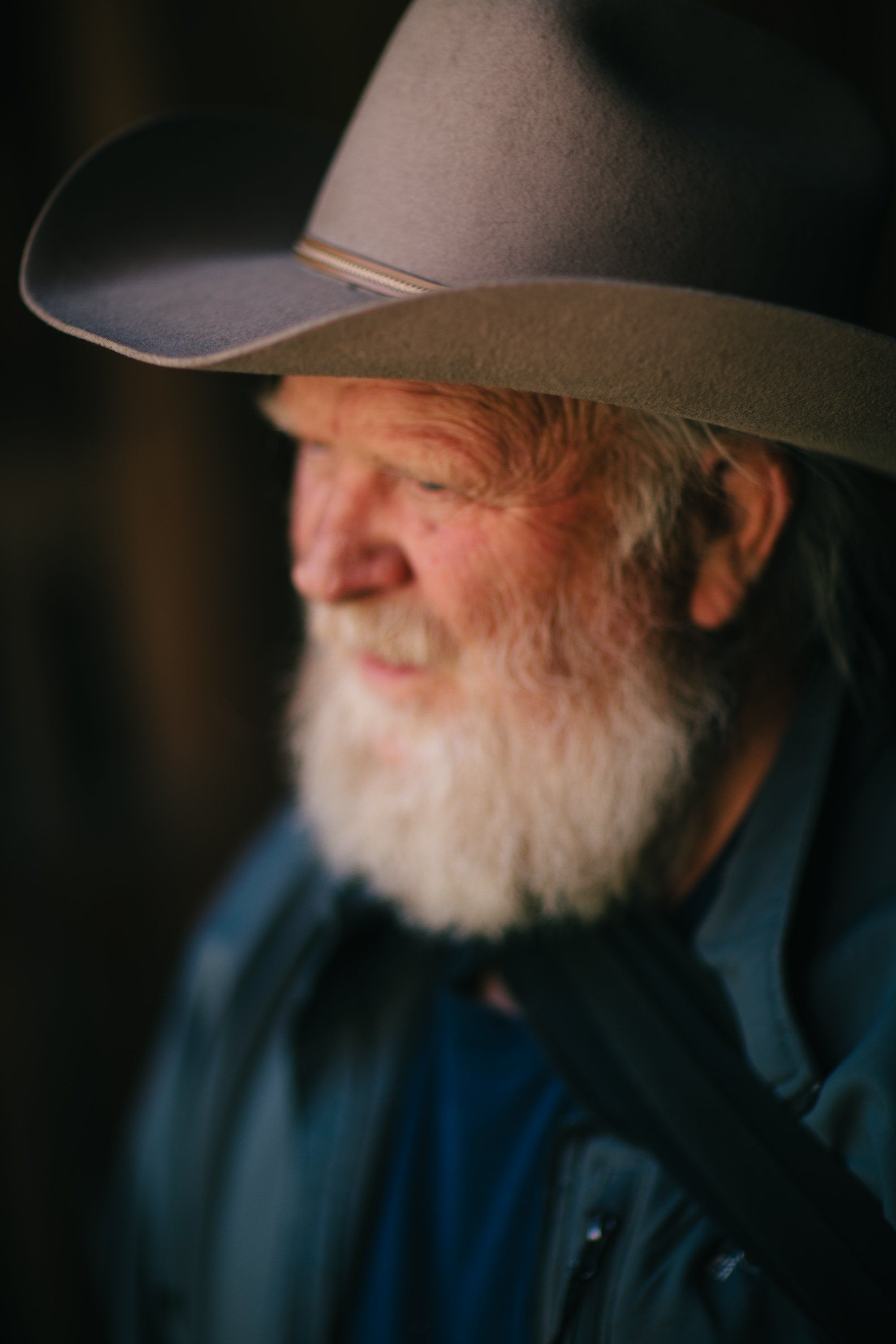 Weathered farmer with white beard in cowboy hat and blue shirt, portrait lit against dark background