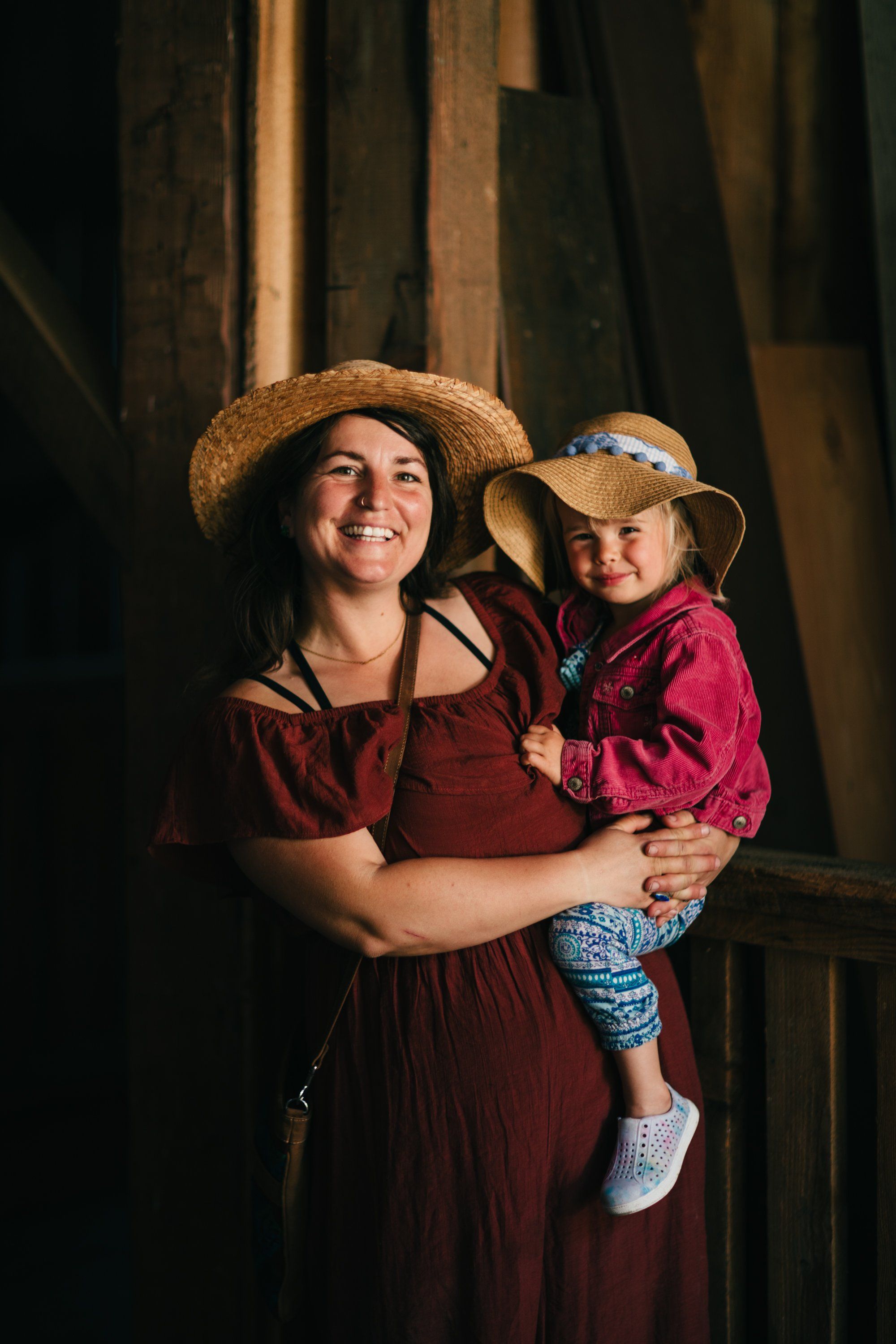 Woman and young child in matching burgundy outfits and straw hats smile in warm sunlight inside a rustic barn