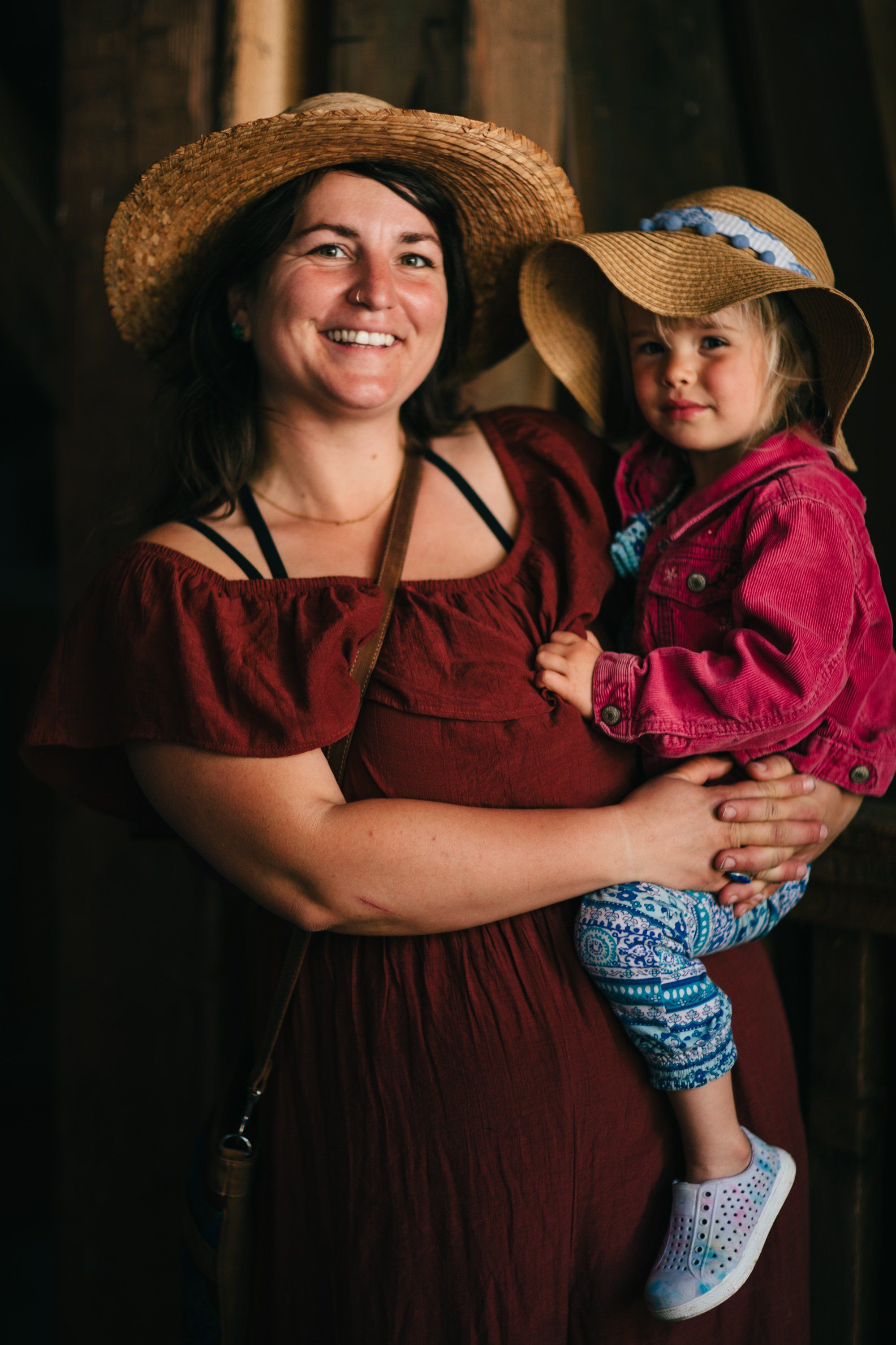 Woman in burgundy dress and straw hat holds smiling toddler in pink jacket against dark barn backdrop