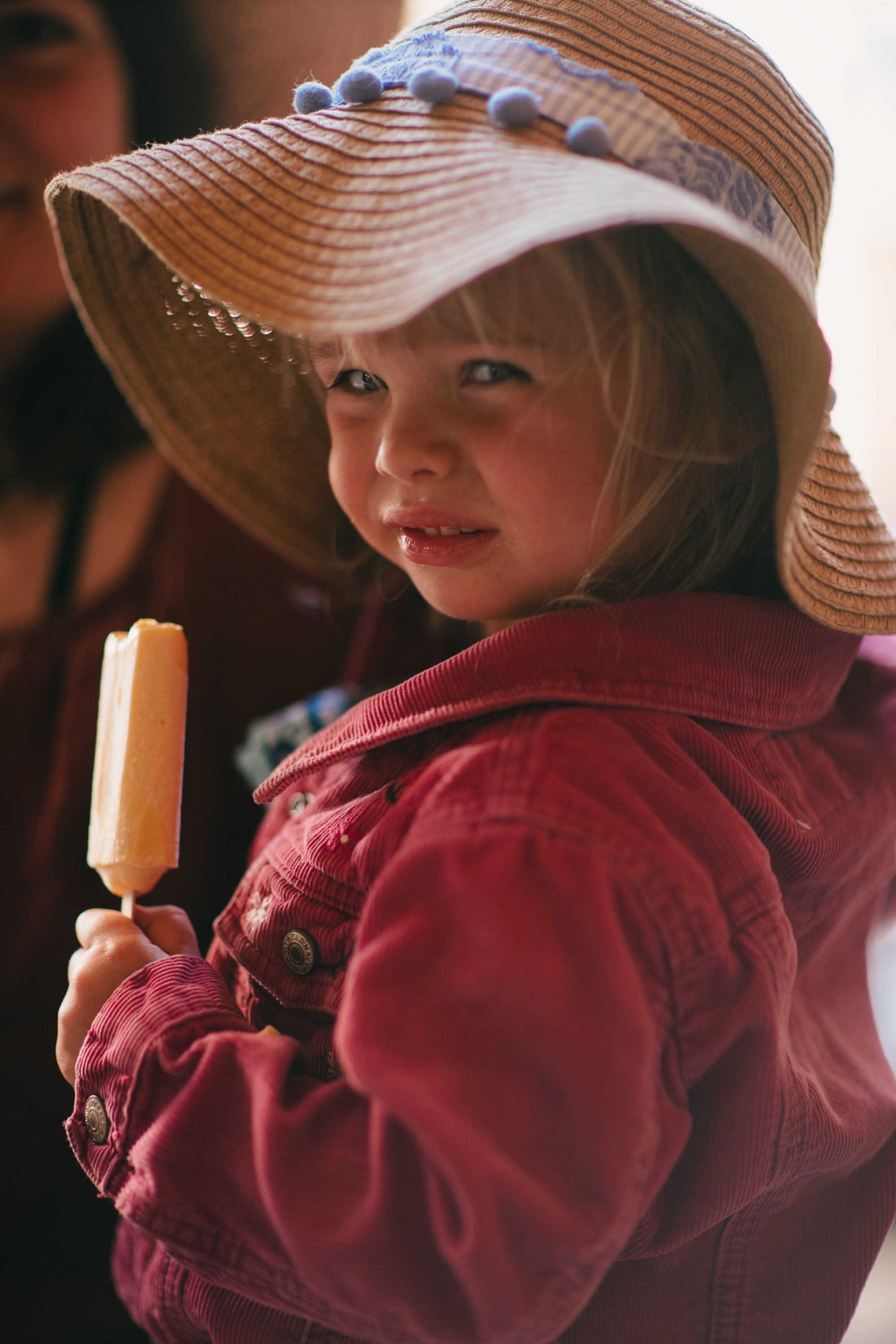 Young child in wide-brimmed straw hat and burgundy jacket holding popsicle, warm golden light, gentle smile toward camera
