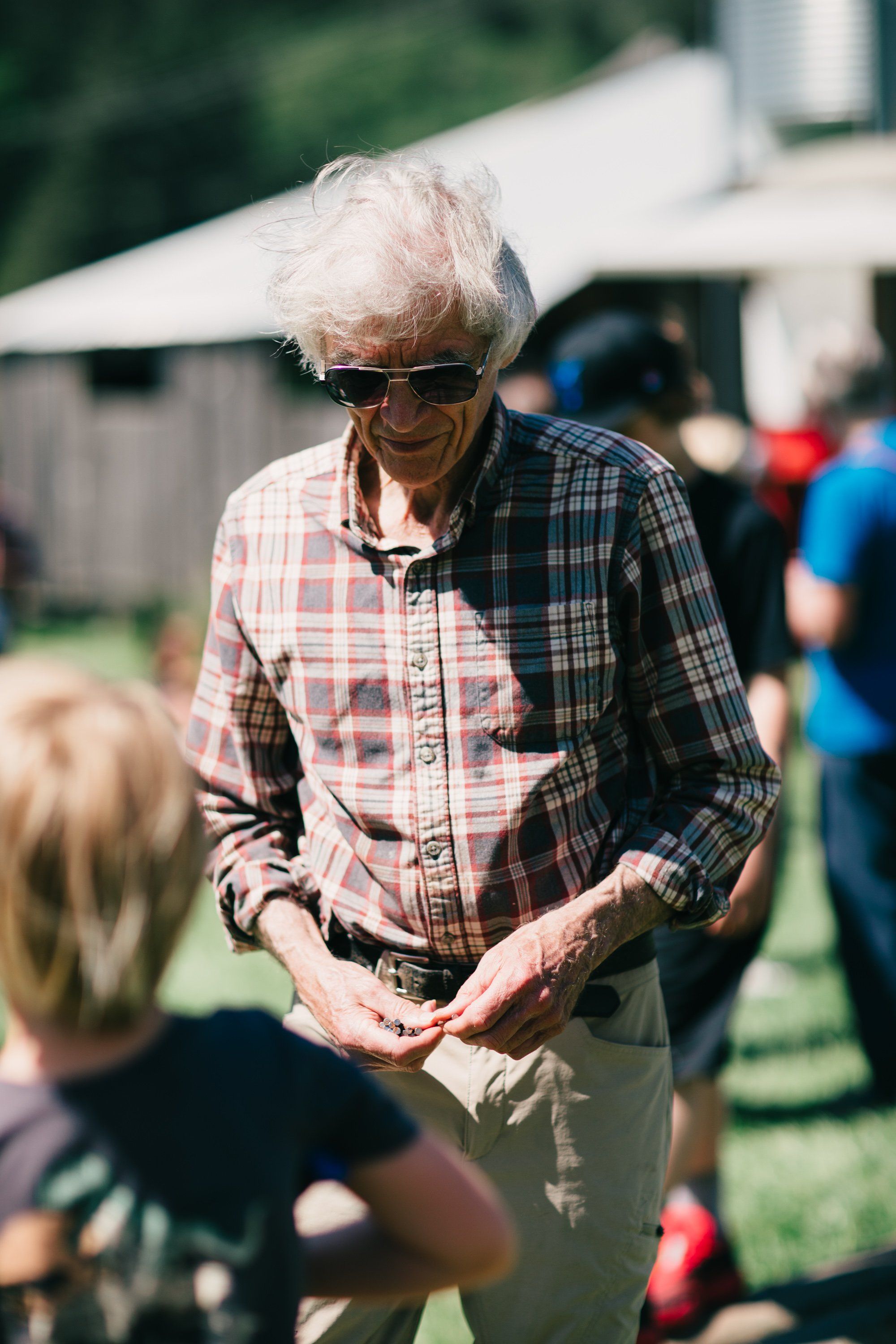 Elderly man in plaid shirt and sunglasses interacts with a child at a sunny farm gathering