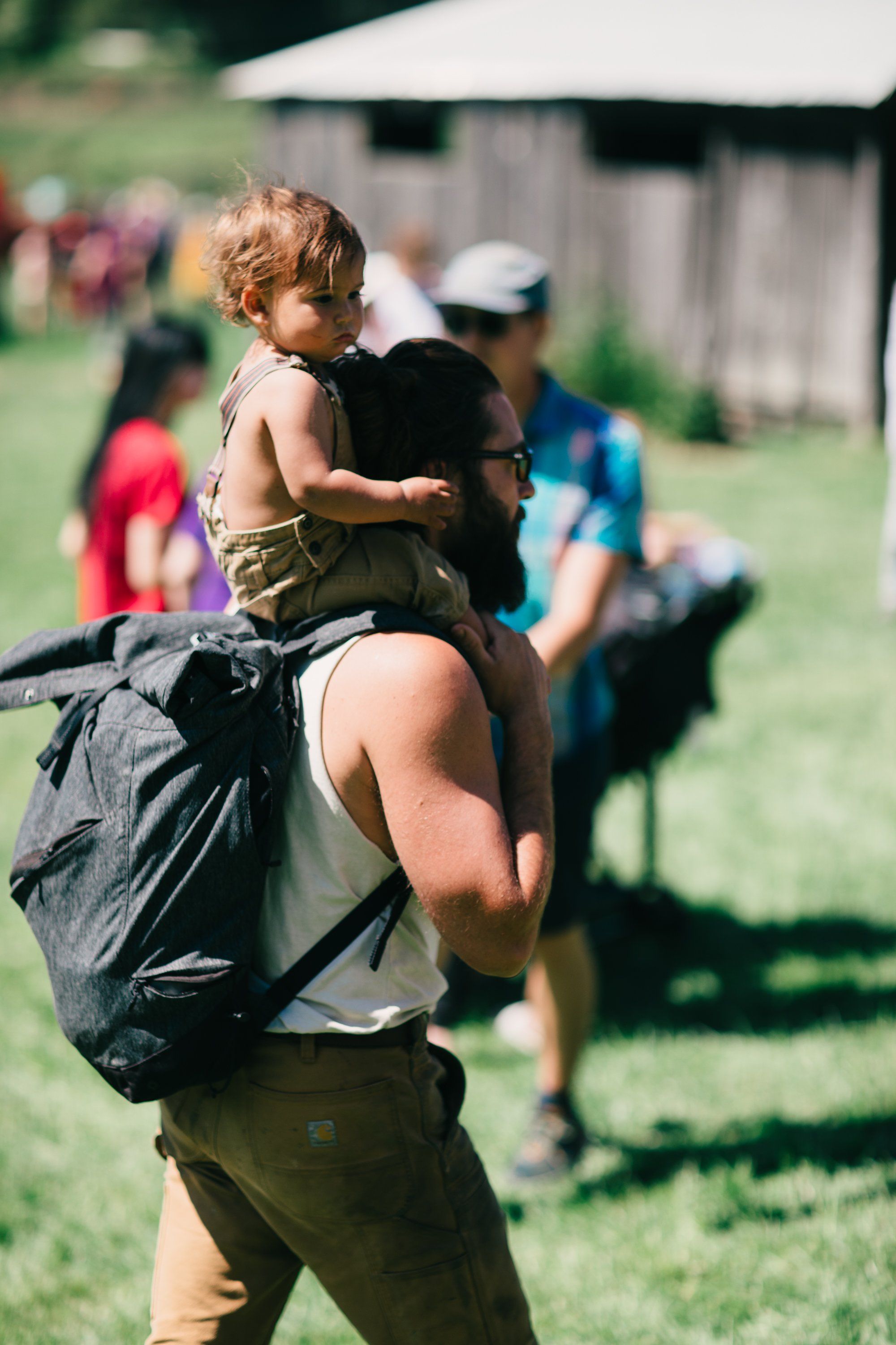 Woman carrying toddler on shoulders at sunny farm gathering, blurred figures and barn in background