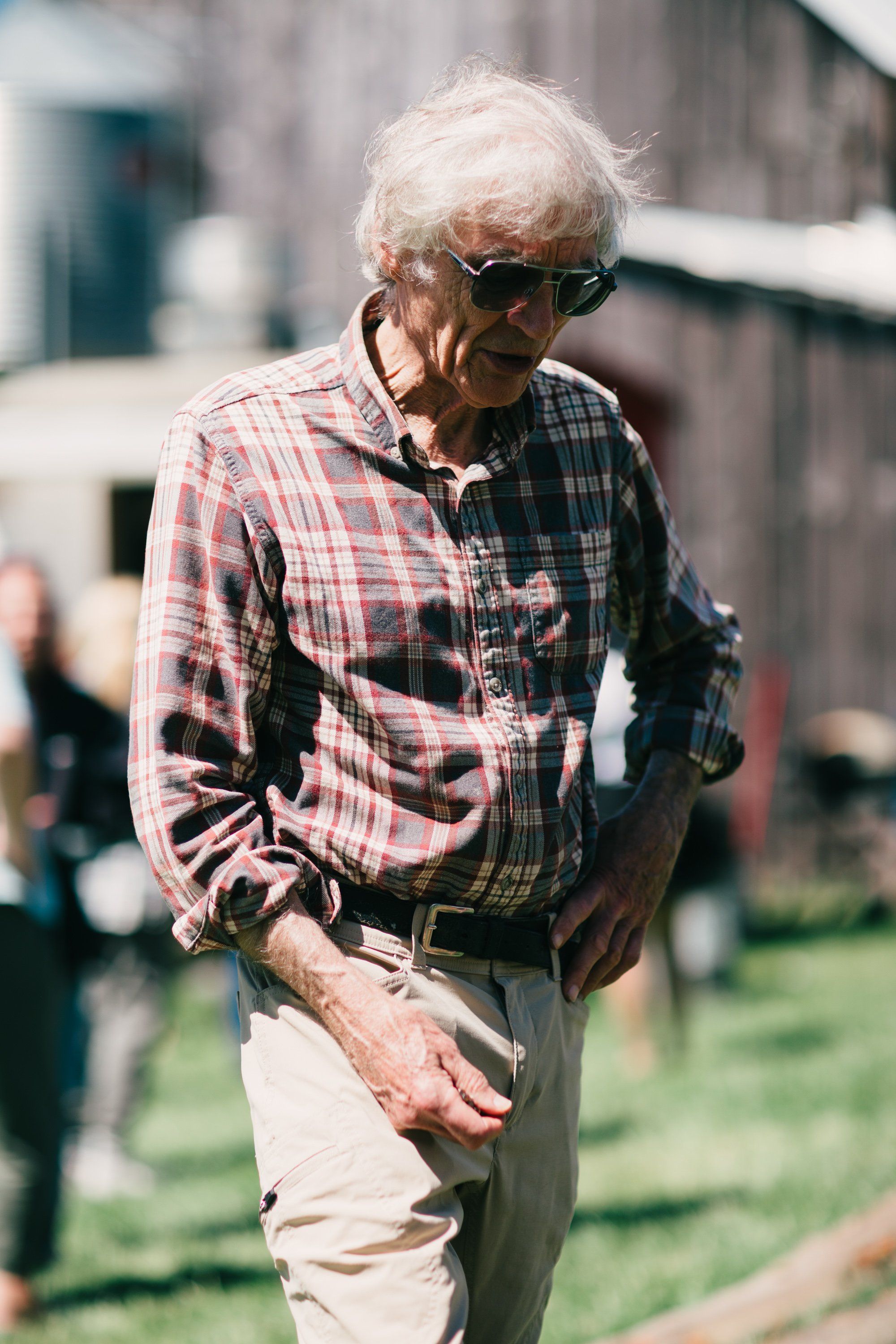 Elderly man in plaid shirt and sunglasses walking through sunny farmyard, soft-focus barn and figures in background