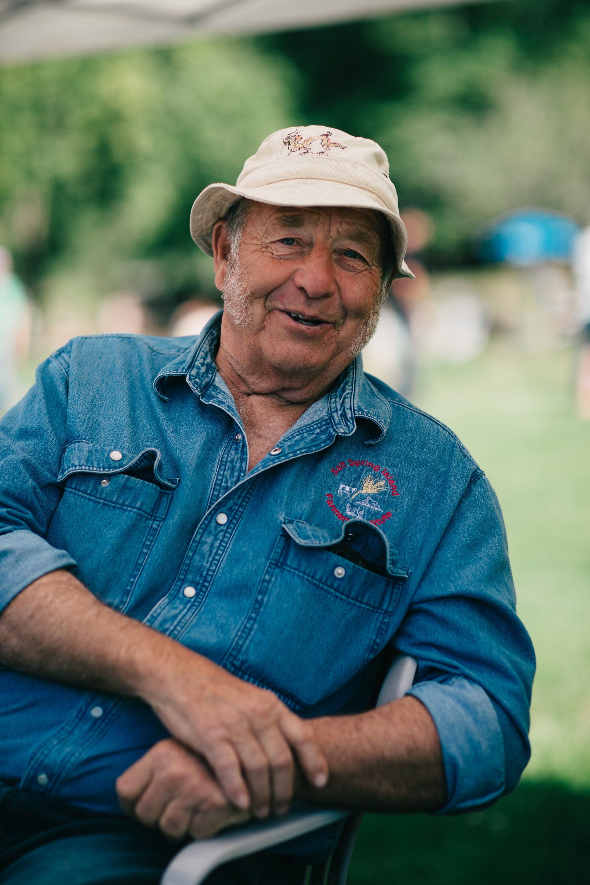 Weathered farmer in denim shirt and cream cap smiles at camera with green fields and farm buildings behind him