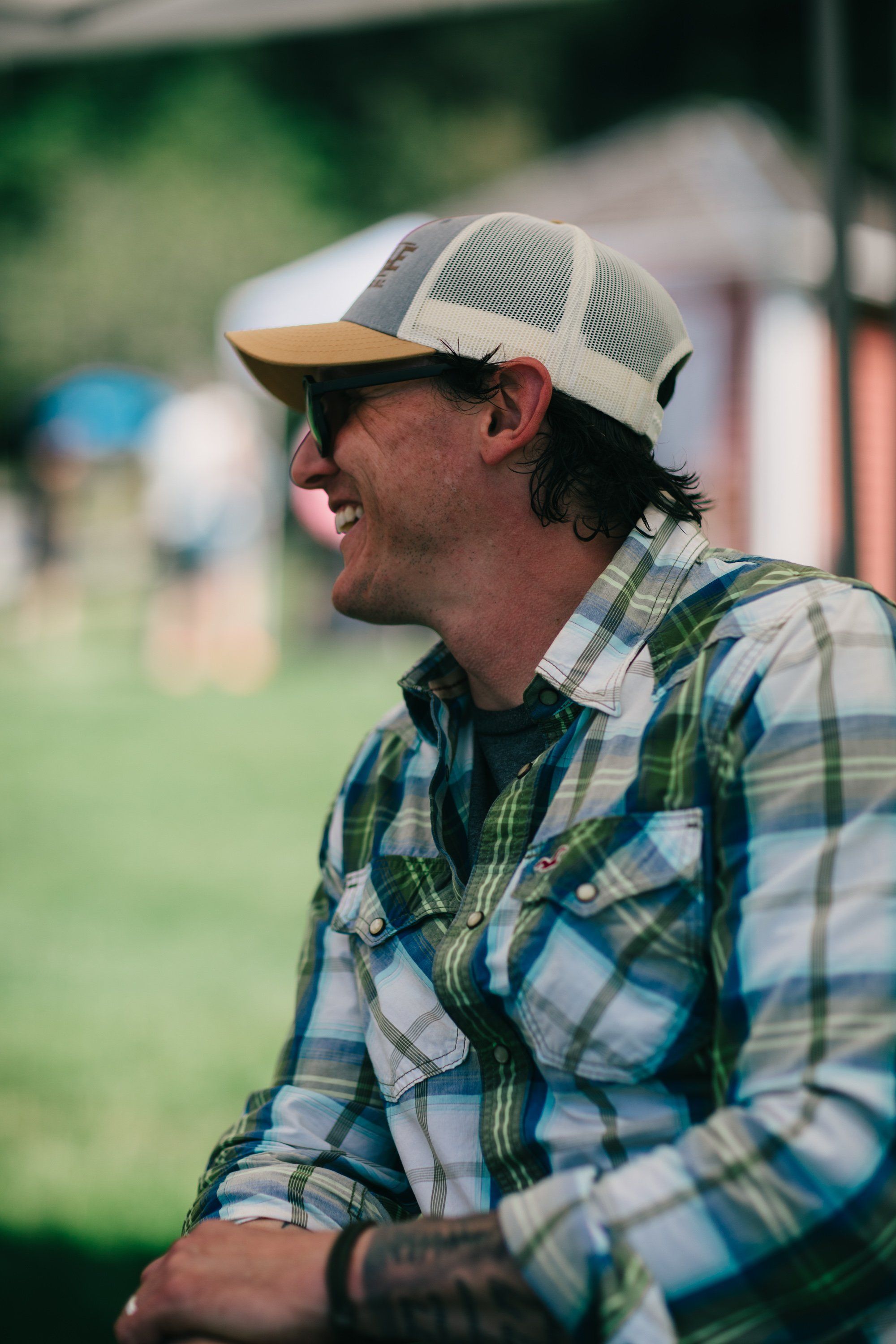 Man in plaid shirt and mesh cap smiles while working outdoors on a sunny farm day, blurred green fields behind him
