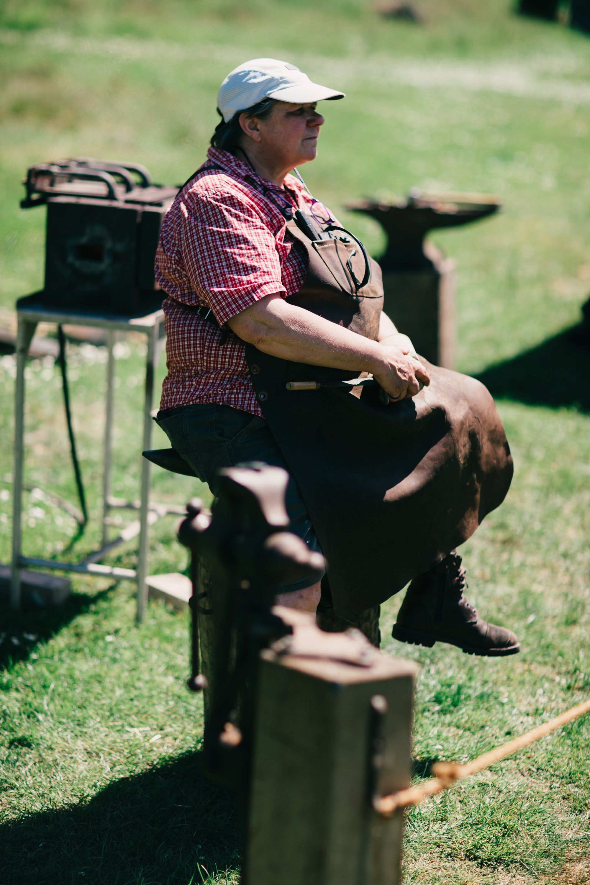 Farmer in red plaid shirt and cap sits on wooden fence post in sunny pasture, hands resting on legs, looking contemplative