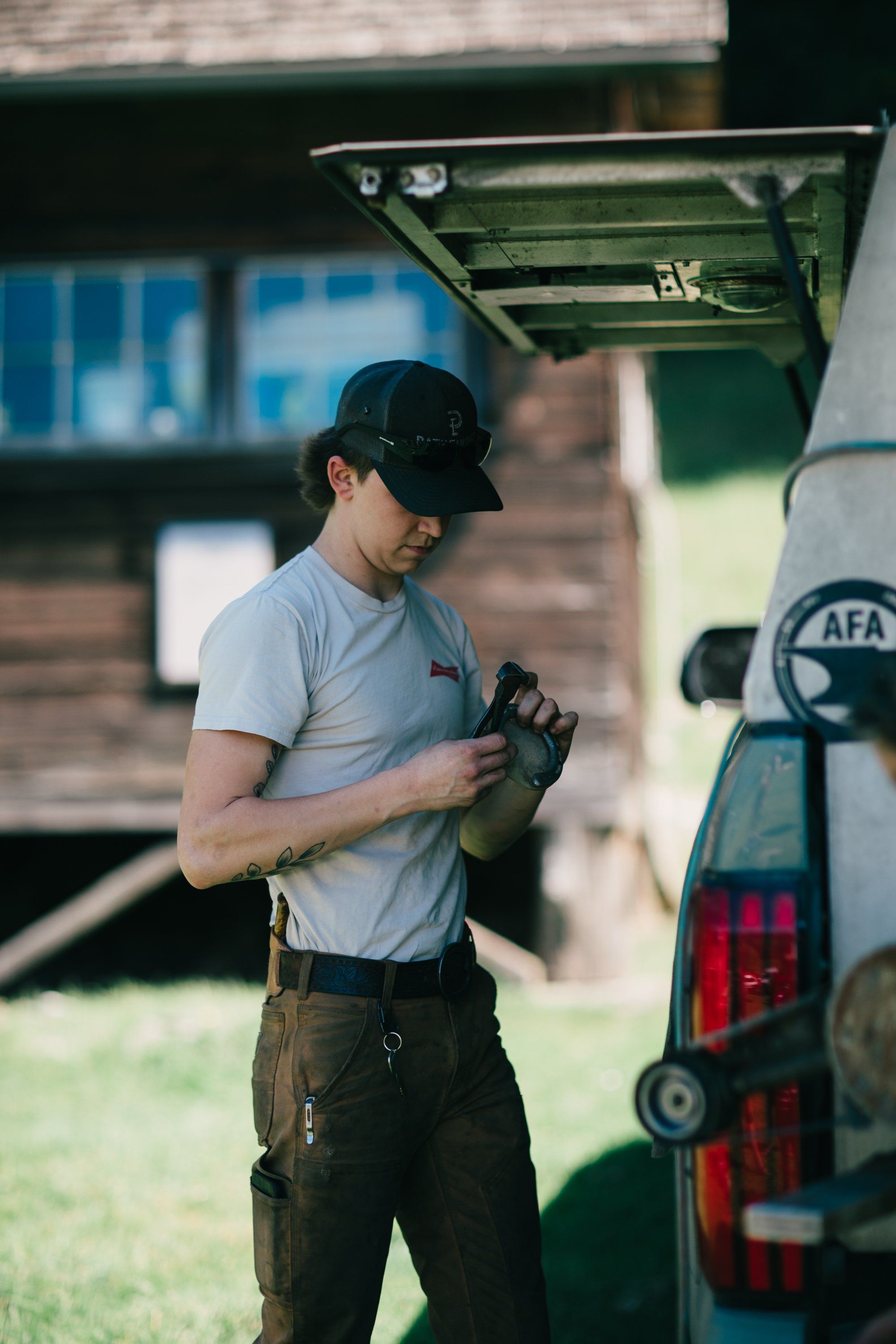 Farmer in grey shirt and cap checks equipment under green awning, golden afternoon light casting shadows across rural workspace