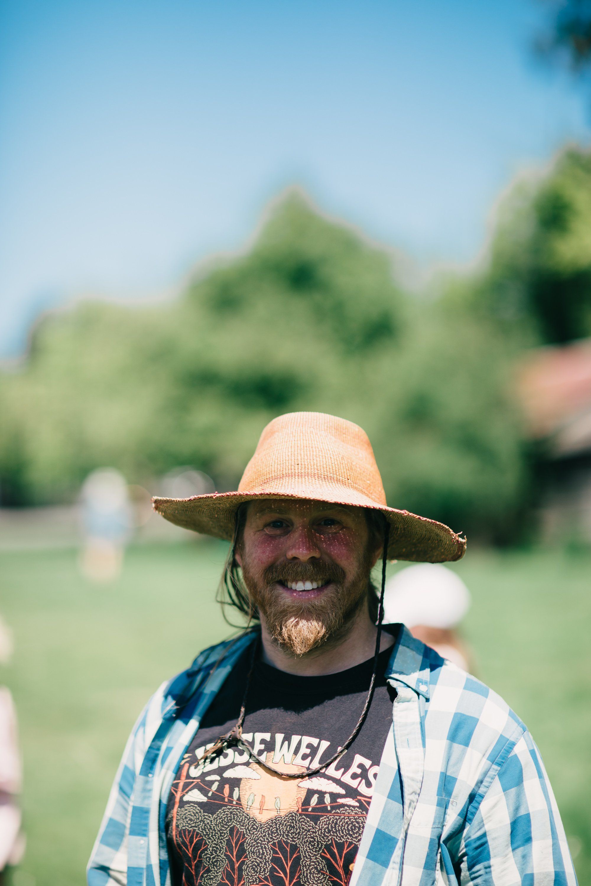 Man in straw hat and blue plaid shirt smiling at camera in sunlit farm field with blurred green trees behind
