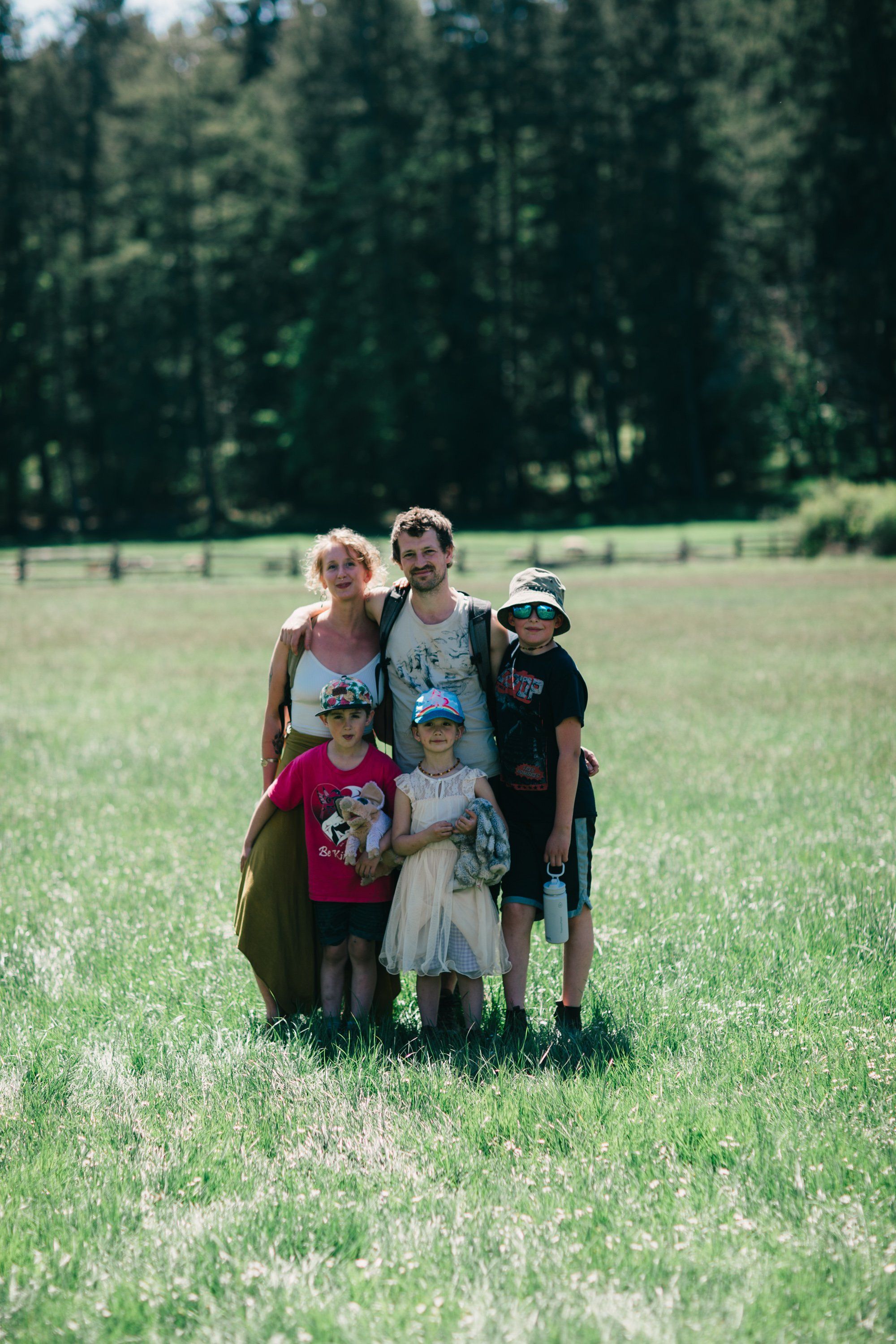 Family of five standing in a sun-dappled meadow with tall evergreen forest backdrop, wearing casual summer clothing