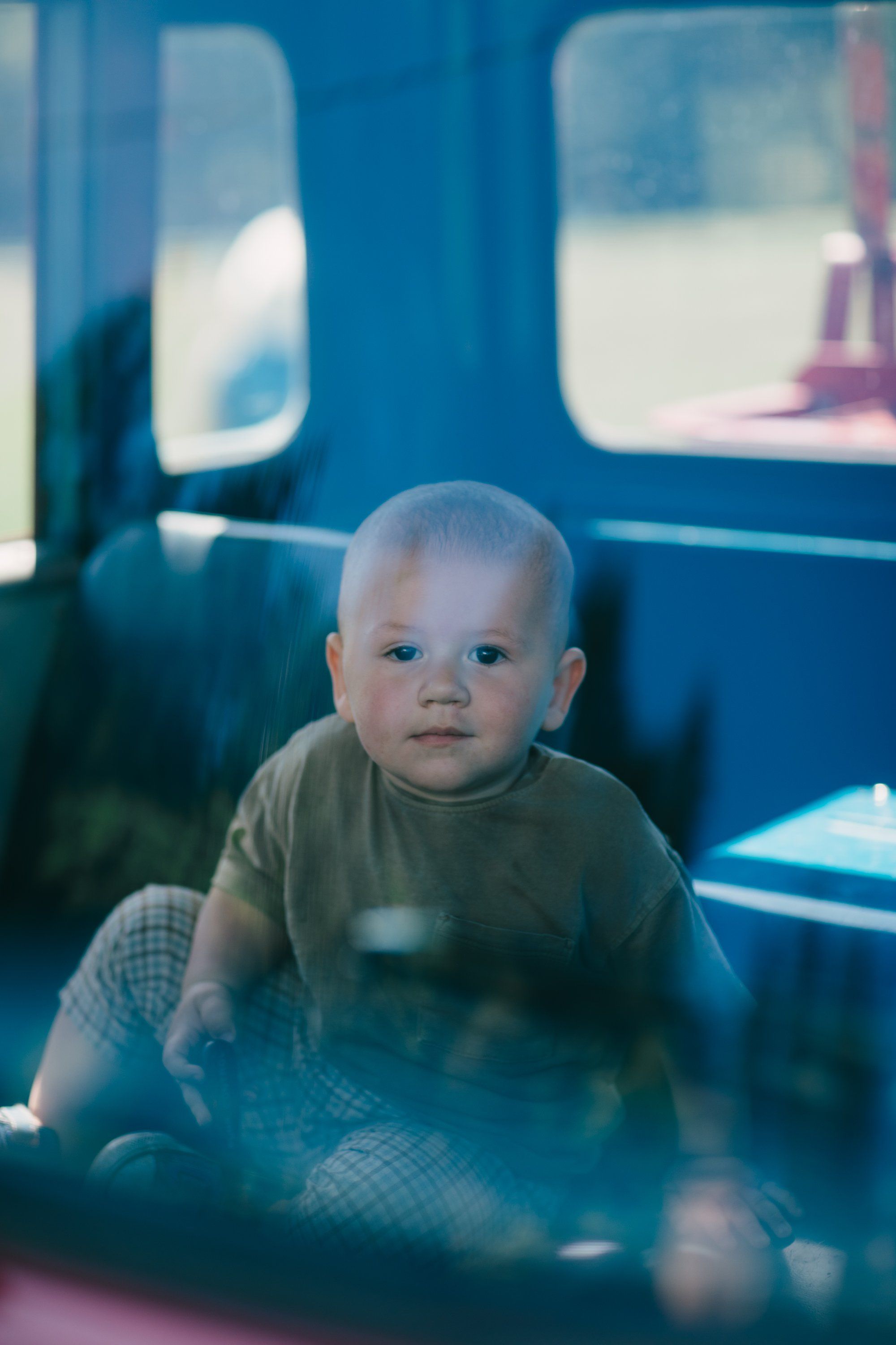 Toddler in olive shirt sitting inside a blue farm vehicle, soft natural light filtering through windows