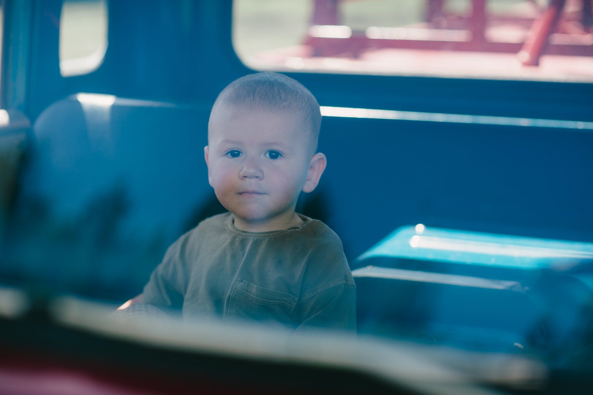 Toddler in soft gray shirt gazes thoughtfully from inside a blue vintage truck cab, warm natural light casting gentle shadows
