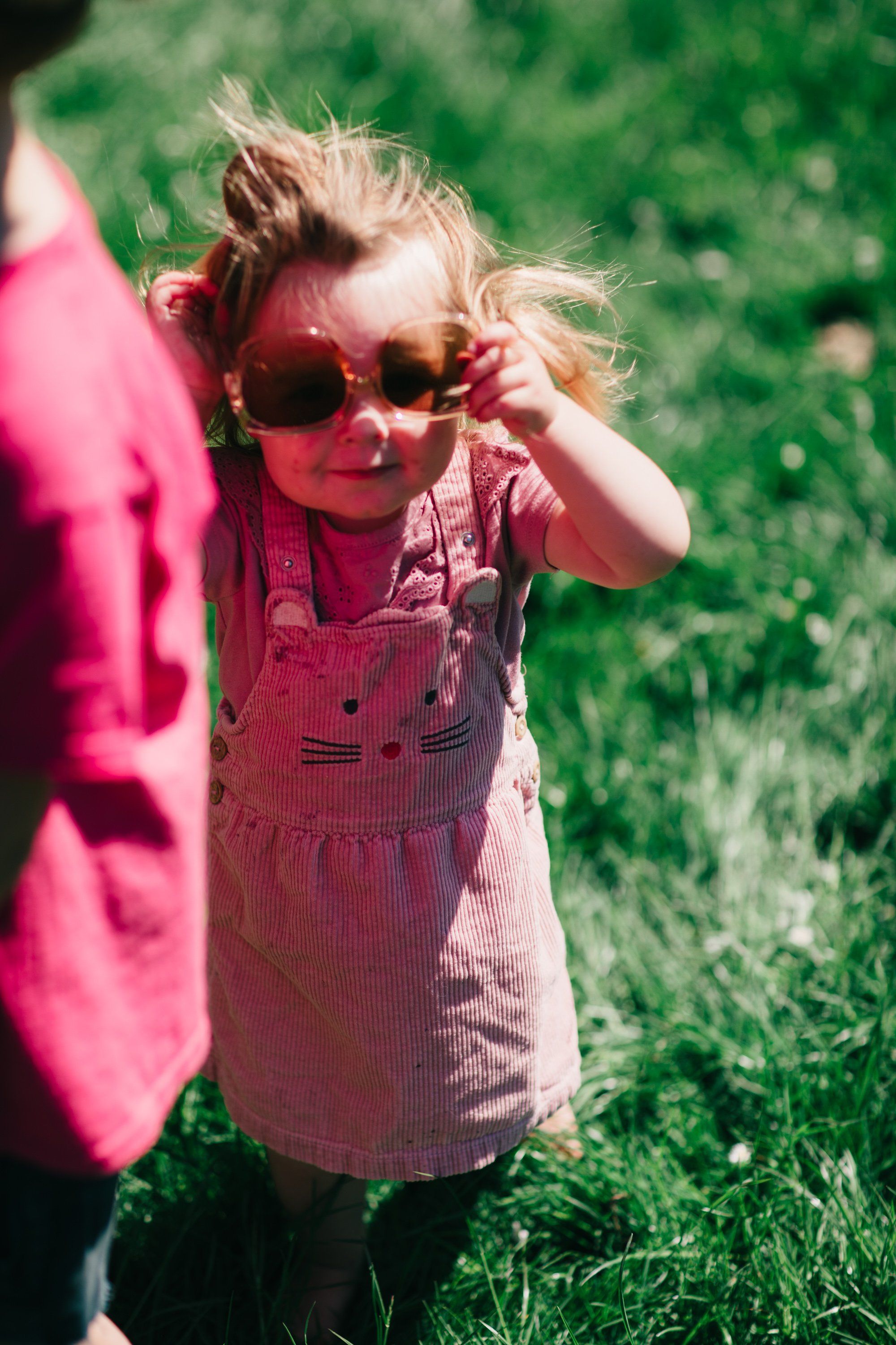 Blonde toddler in pink cat-print dress and oversized sunglasses stands in sunny grass, hand raised to adjust glasses