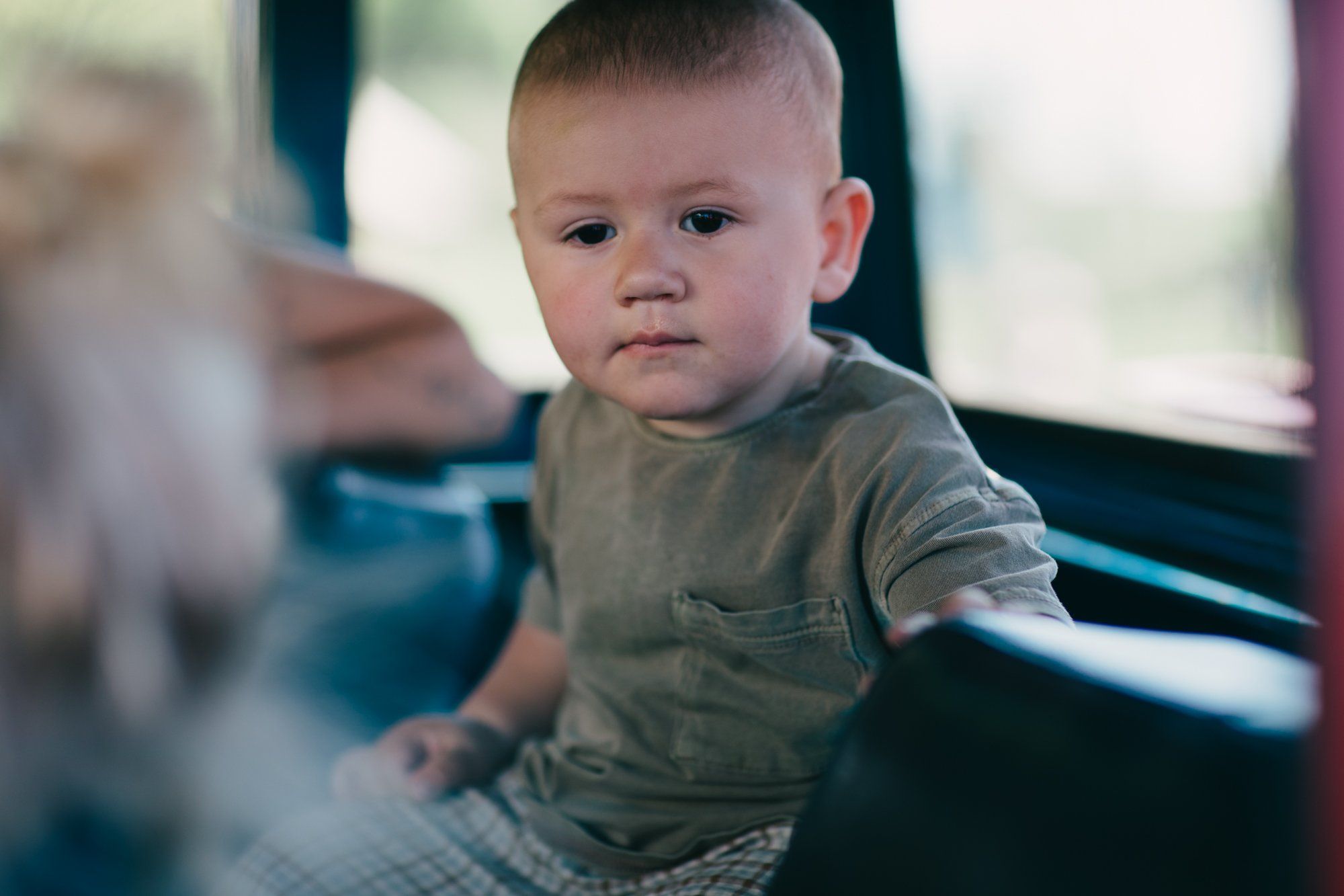 Toddler in olive shirt sits in soft natural light, gazing thoughtfully while an adult's hand rests nearby in a rustic farm setting