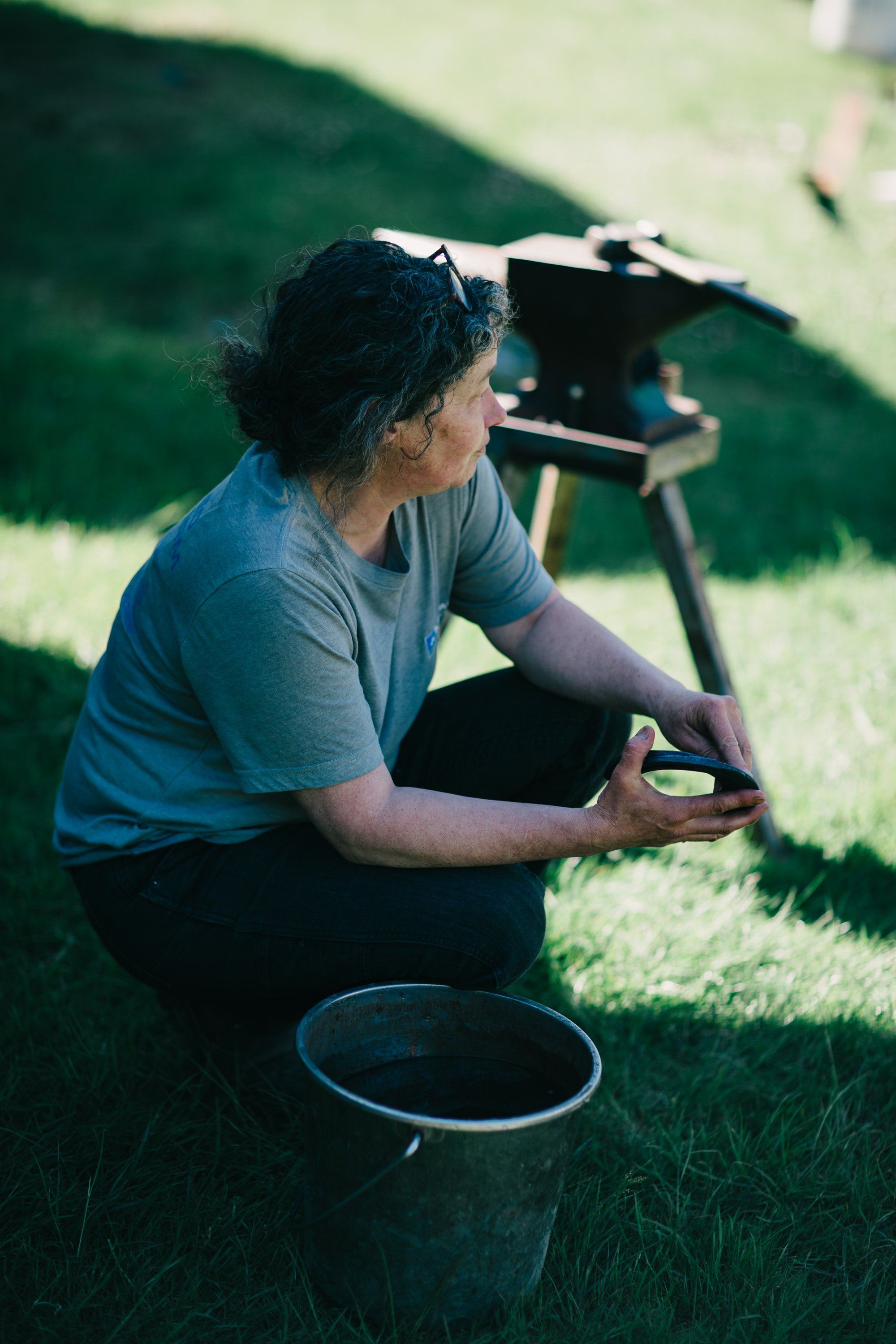 Photographer crouches in sunlit grass with camera on tripod, holding light meter, soft afternoon light casting gentle shadows across the field