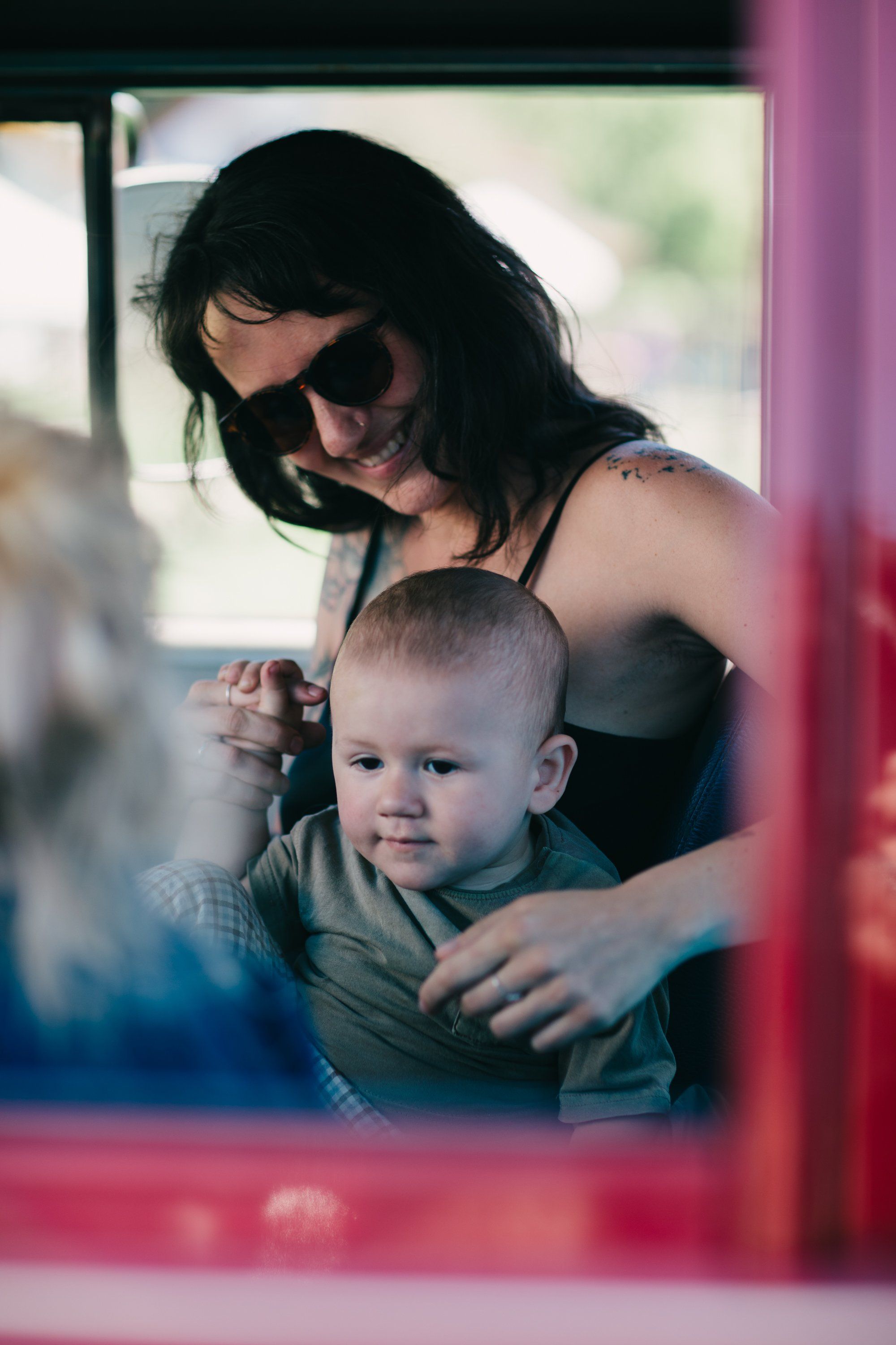 Woman in sunglasses holding a smiling toddler by a bright pink window frame, soft natural light
