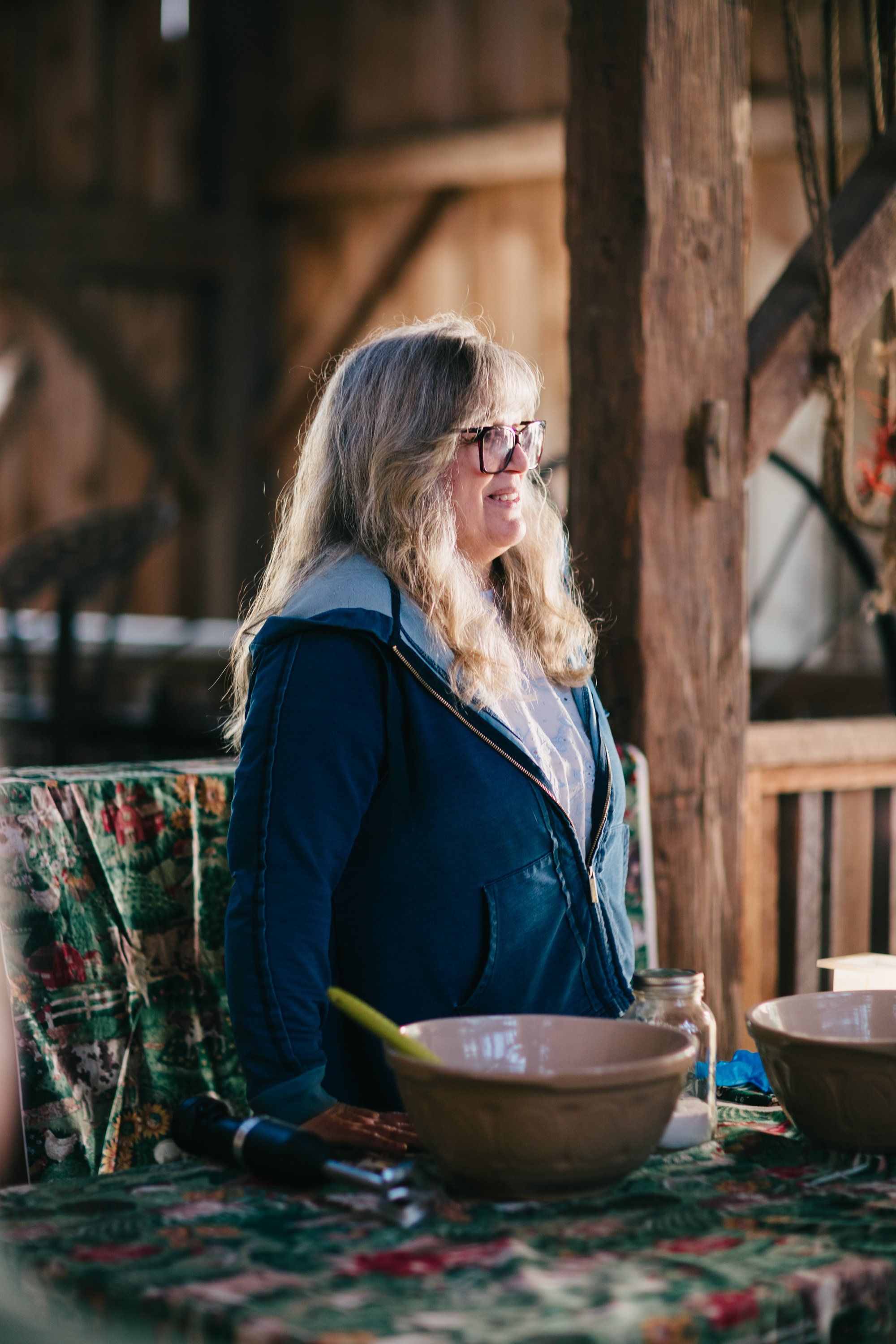 Woman with long gray hair and glasses in navy jacket sits thoughtfully beside wooden bowls in a rustic farm setting
