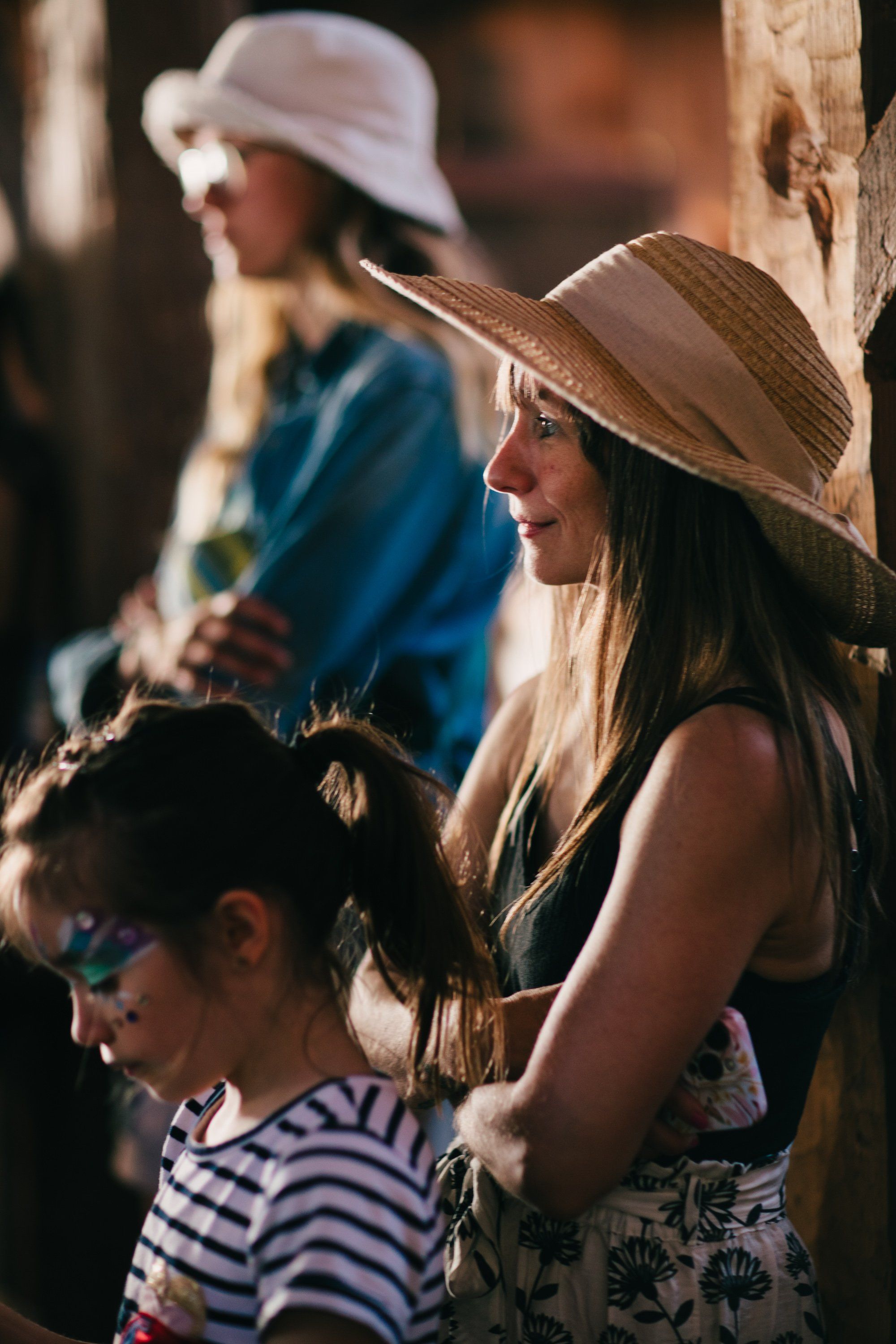 Woman in straw hat holding young child, standing beside wooden fence in warm afternoon light, with blurred figures in background