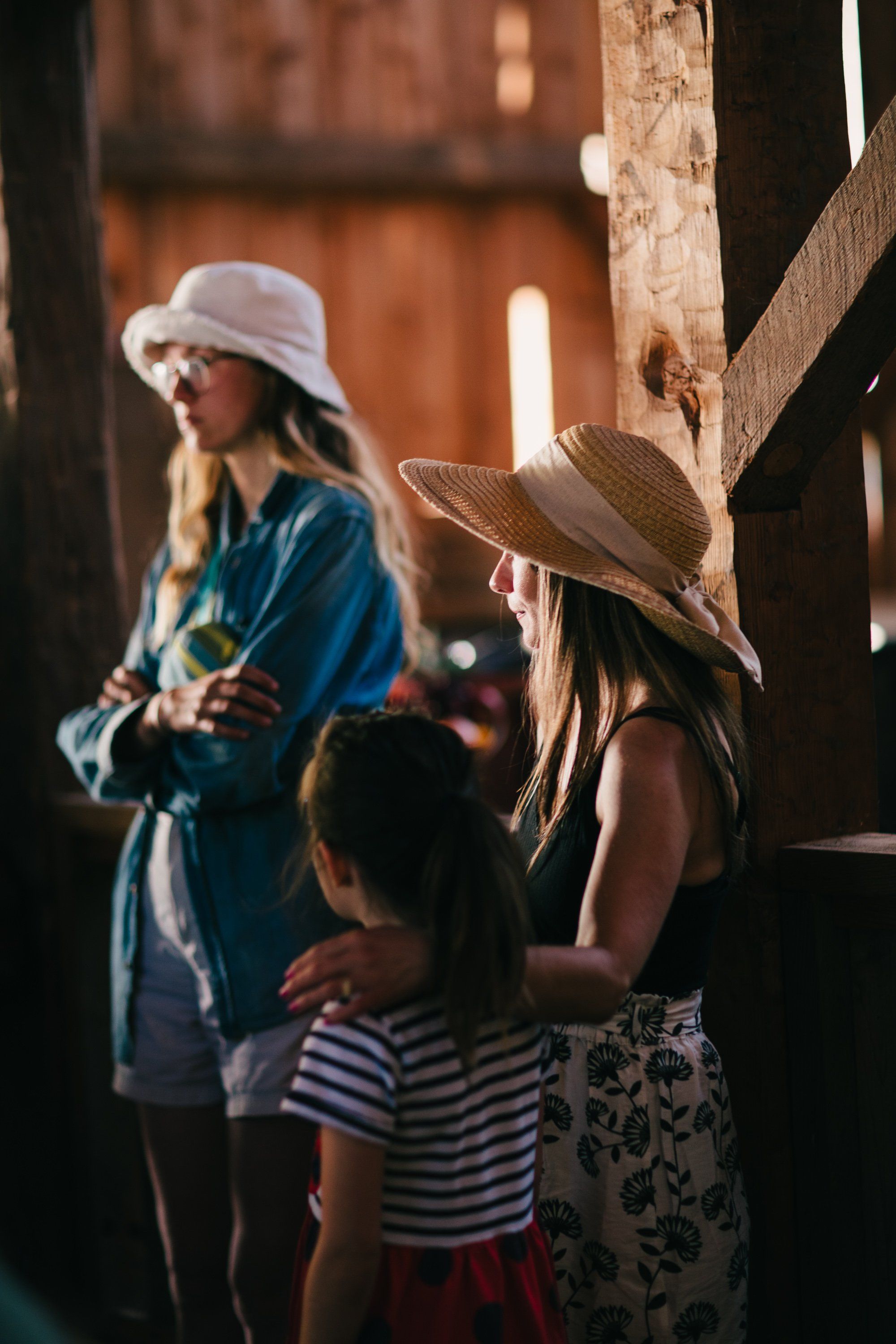 Two women in wide-brimmed hats and a child in striped shirt stand together in warm golden light within a rustic wooden structure