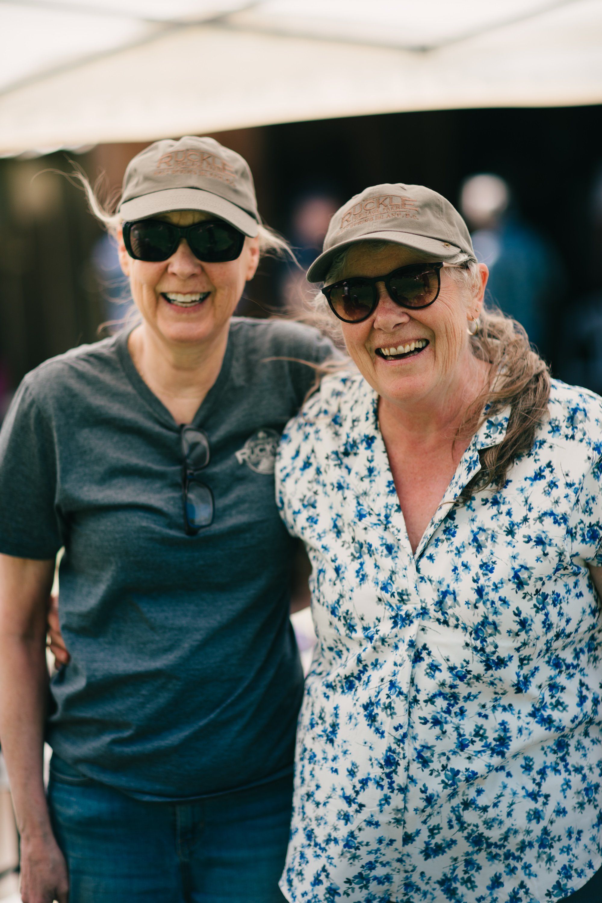 Two smiling women wearing caps and sunglasses stand together under tent shade at a farm event
