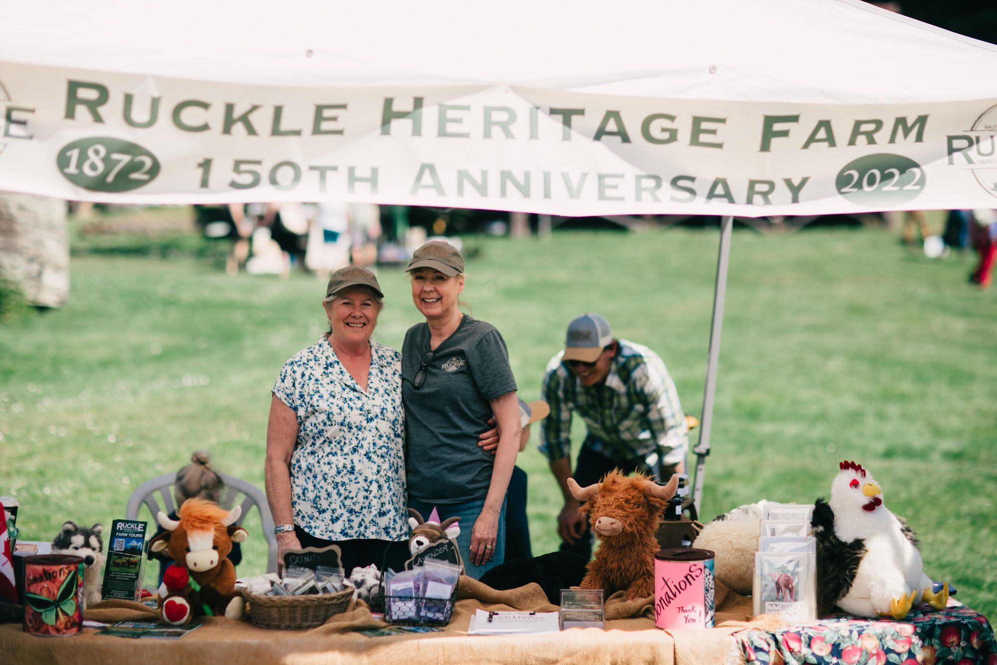 Three people stand behind a table displaying farm goods under a banner celebrating Ruckle Heritage Farm's 150th anniversary, with green fields and vintage to…