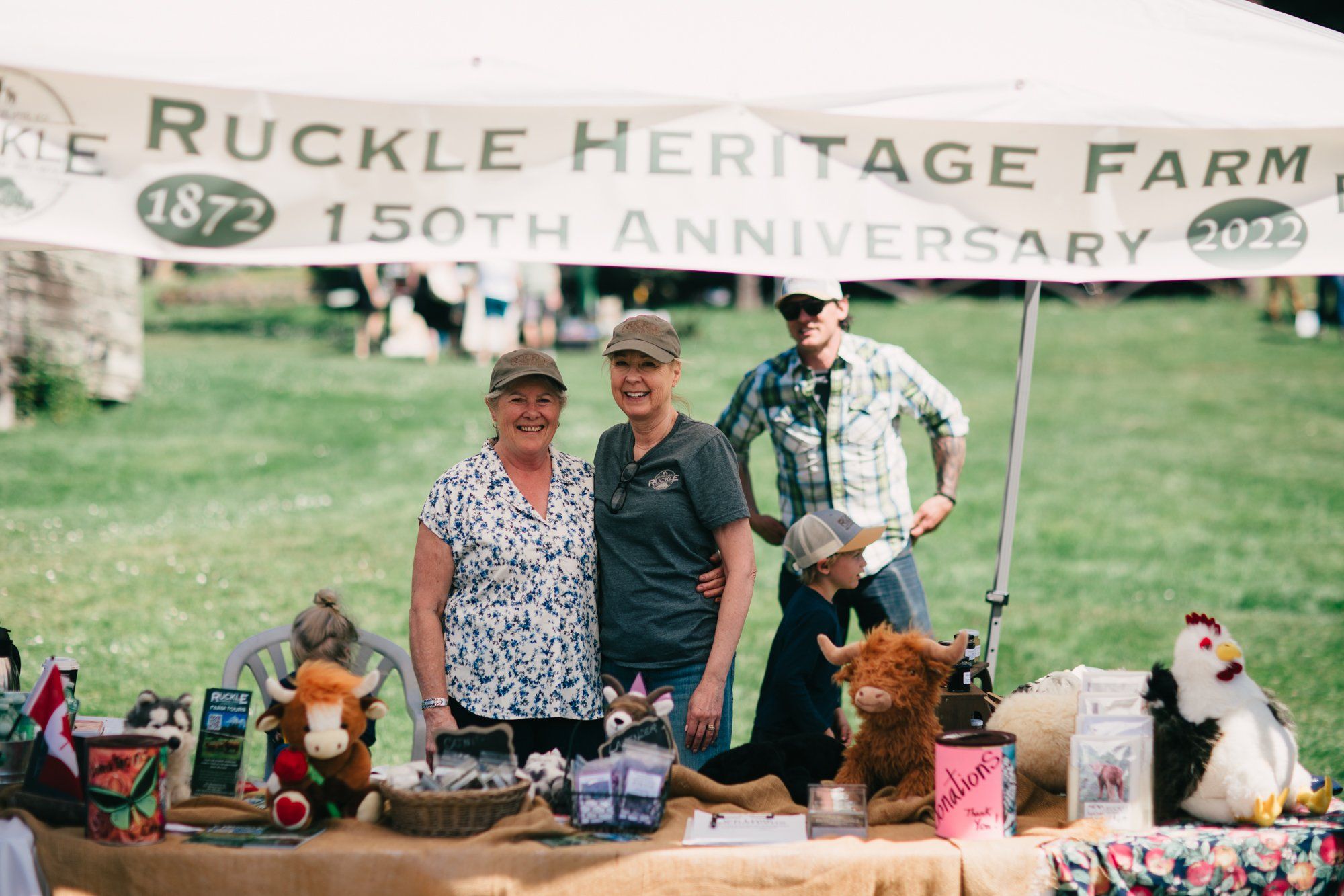 Three people stand behind a farm stand display at Ruckle Heritage Farm's 150th anniversary celebration, surrounded by crafts and goods