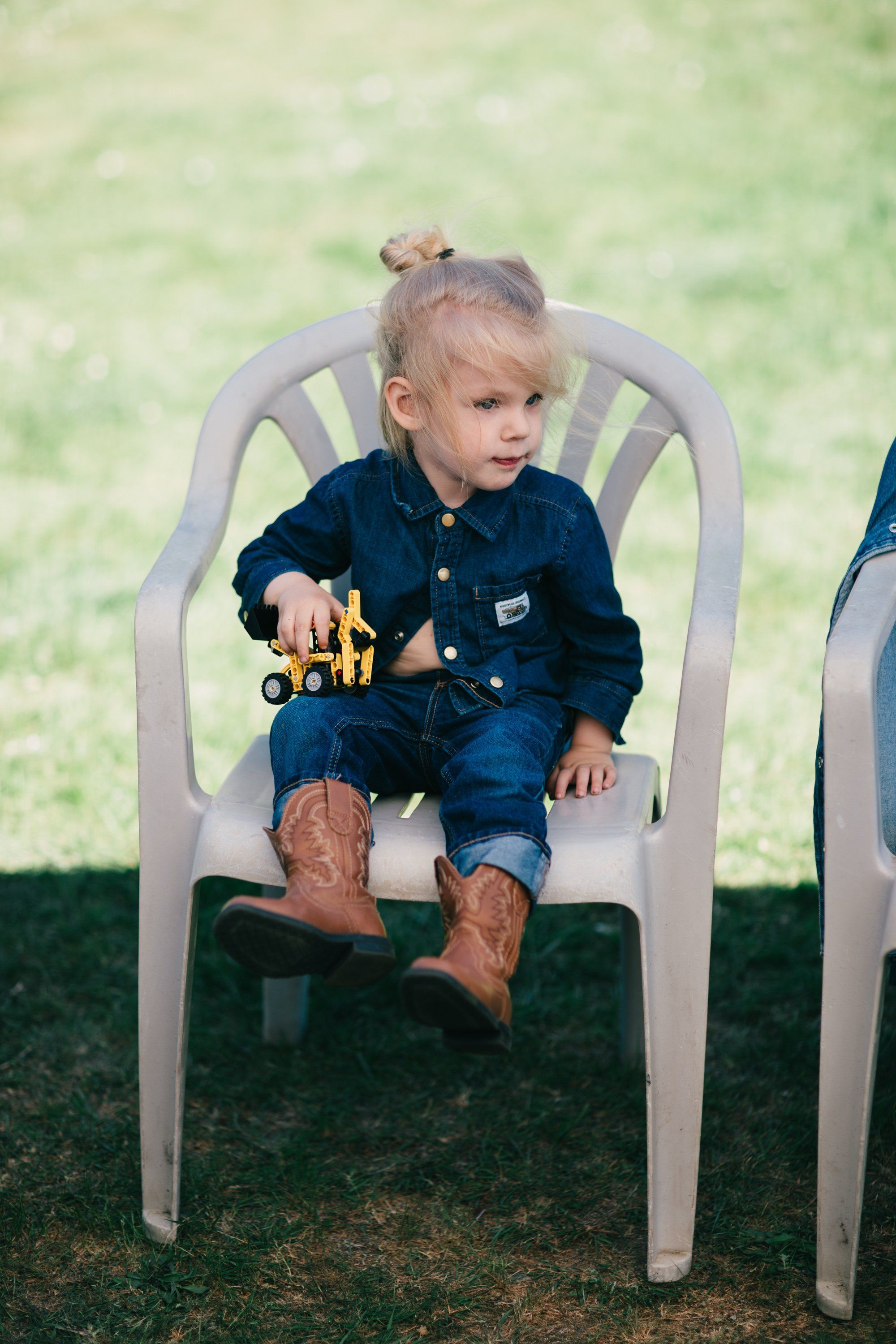Toddler in denim shirt sits on white metal chair in grassy field, holding toy construction equipment, soft afternoon light